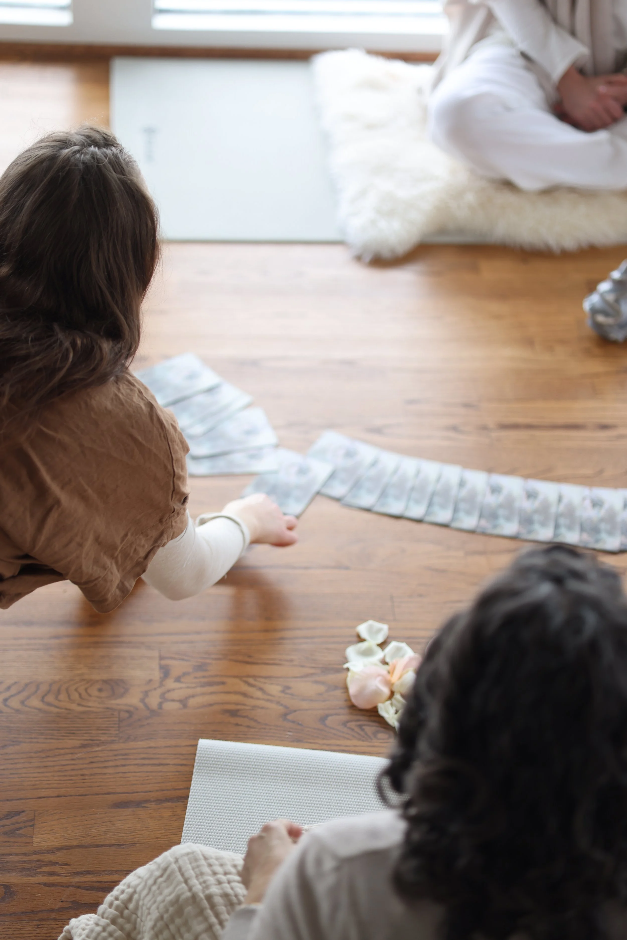 Two women sitting on a wooden floor, one with dark hair in a bun, and the other with curly hair, playing a card game with tarot cards, with candles and flowers nearby, in a cozy indoor setting.