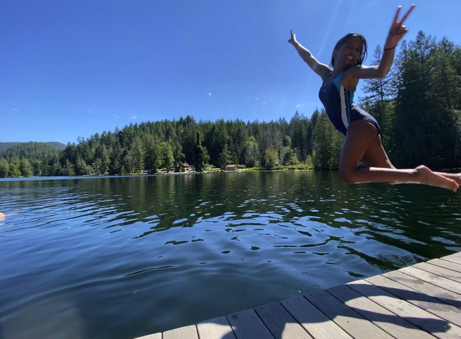 A girl in a swimsuit jumping into a lake with a forested shoreline and clear blue sky in the background.