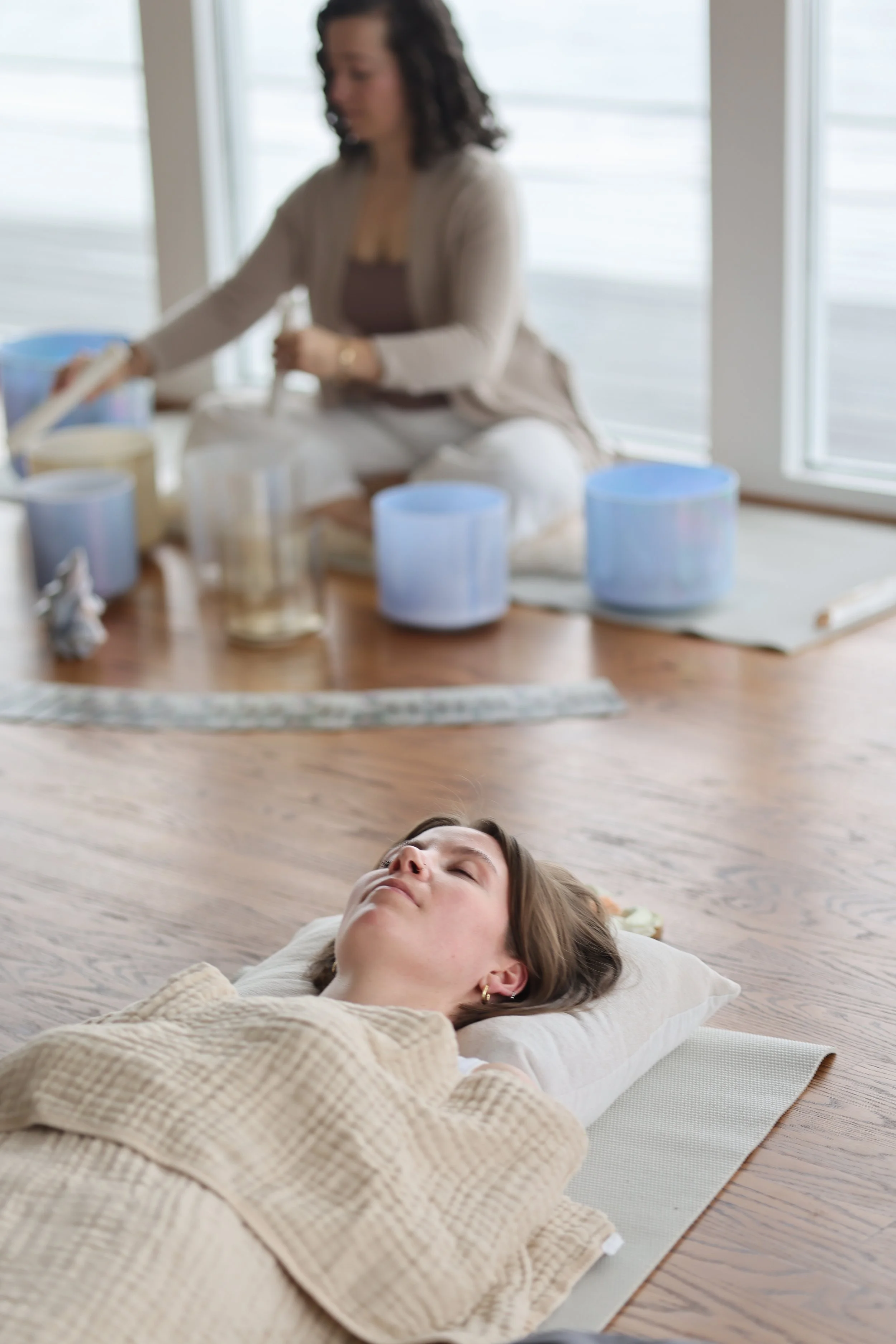 A woman is lying on a pillow on the wooden floor, sleeping or resting. In the background, a woman is sitting on a mat, surrounded by singing bowls and other meditation instruments.