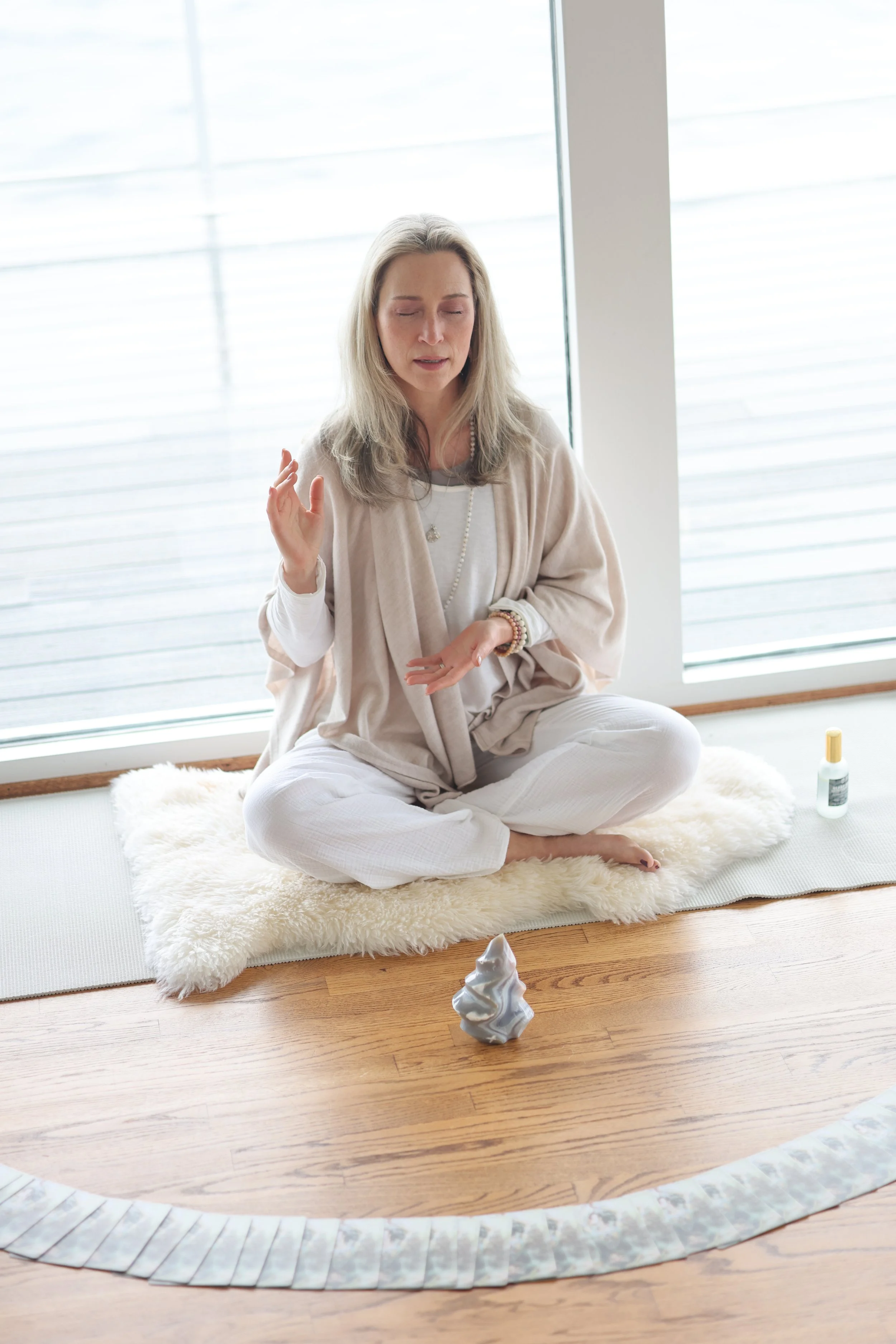 A woman sits cross-legged on a fluffy white rug in front of large windows, with a small bottle of oil nearby. She is raising one hand and appears to be practicing meditation or mindfulness.