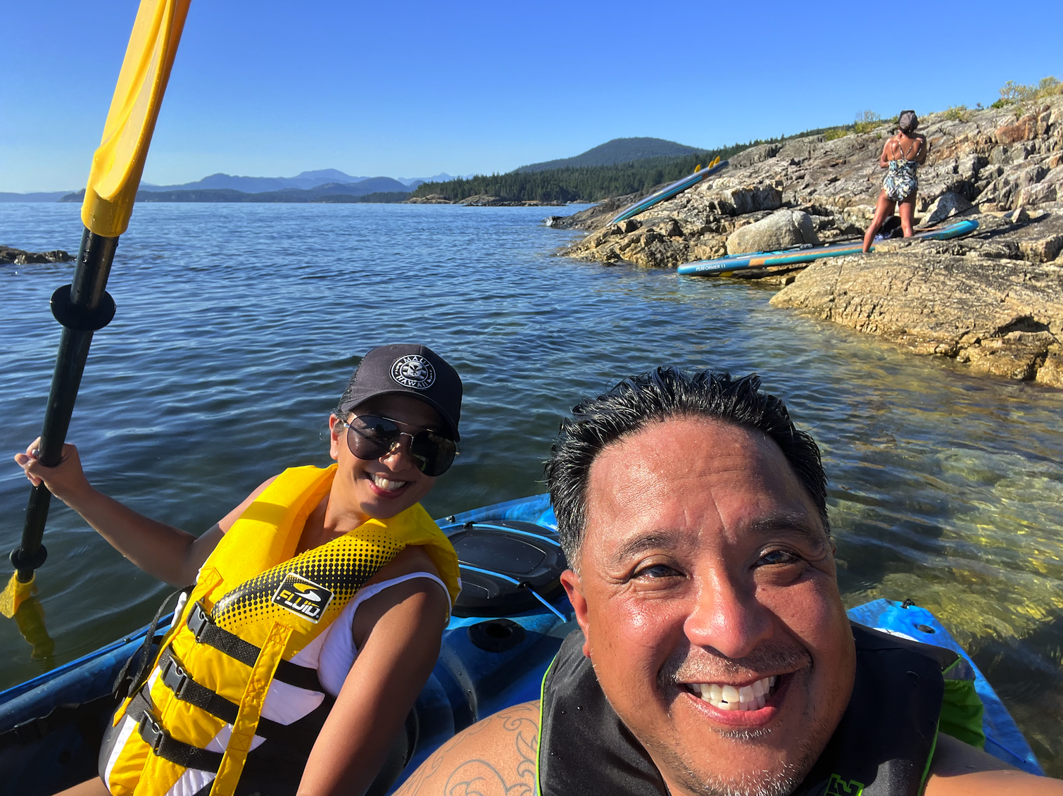 Two people taking a selfie on a kayak near a rocky shoreline with another person in the background preparing to paddle, all in a scenic body of water with mountains and trees in the distance.