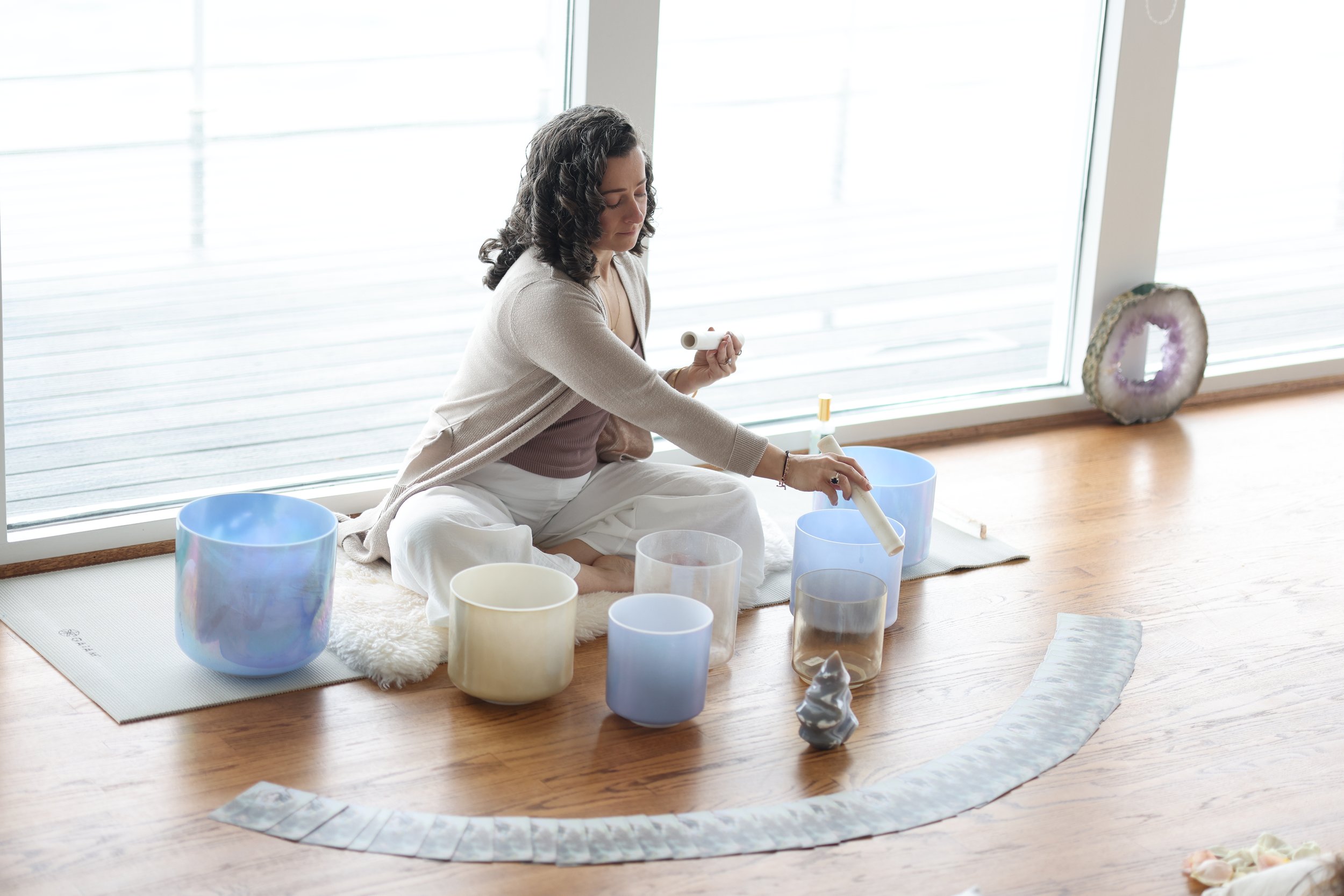 A woman sitting cross-legged on a rug near a large window, playing crystal singing bowls with a mallet in her right hand and a stick in her left hand, surrounded by various other singing bowls and a small ornamental object on the floor.
