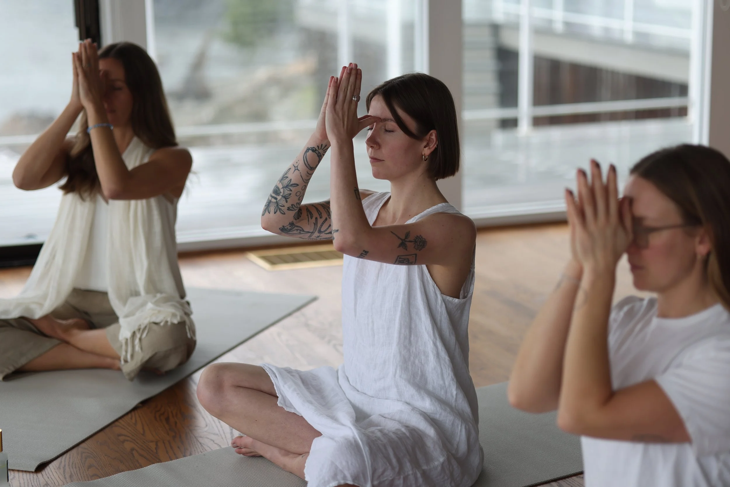 Three women practicing meditation on yoga mats inside a room with large windows, sitting cross-legged with hands pressed together near their foreheads.