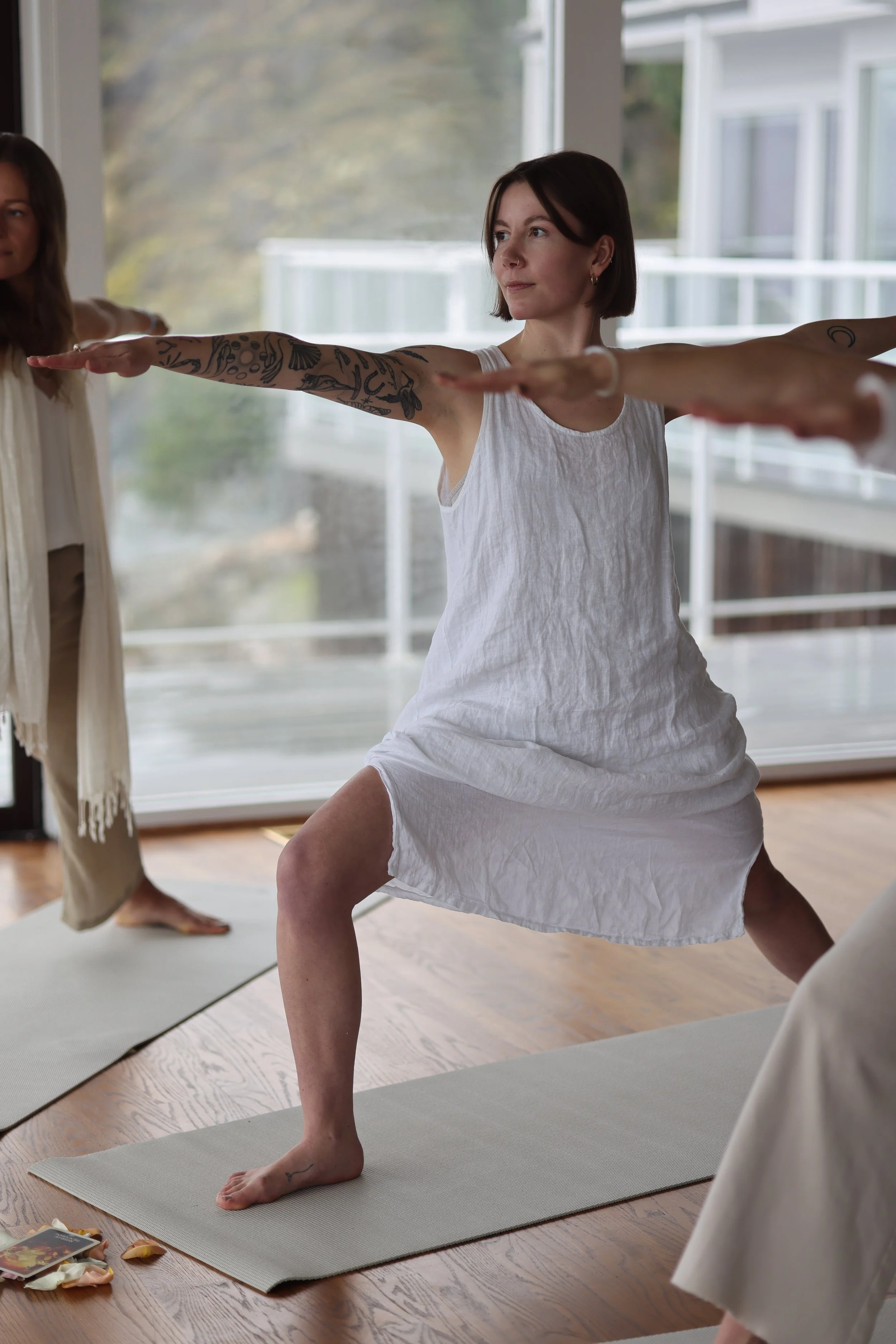 Woman in a white dress practicing yoga with arms extended, in a lunge pose on a yoga mat in a room with large windows.