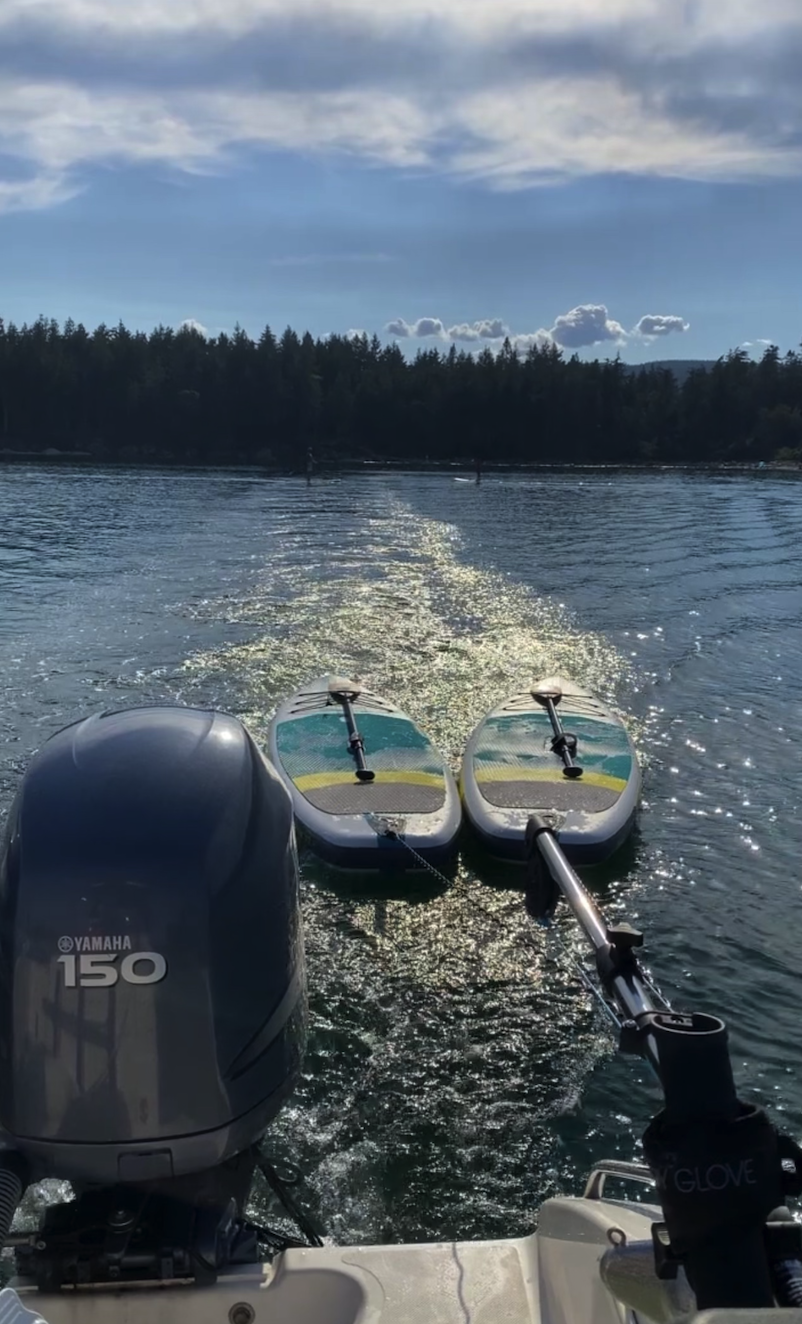 View from a boat showing two paddleboards and a Yamaha 150 outboard motor on a body of water with a forested shoreline and a partly cloudy sky.