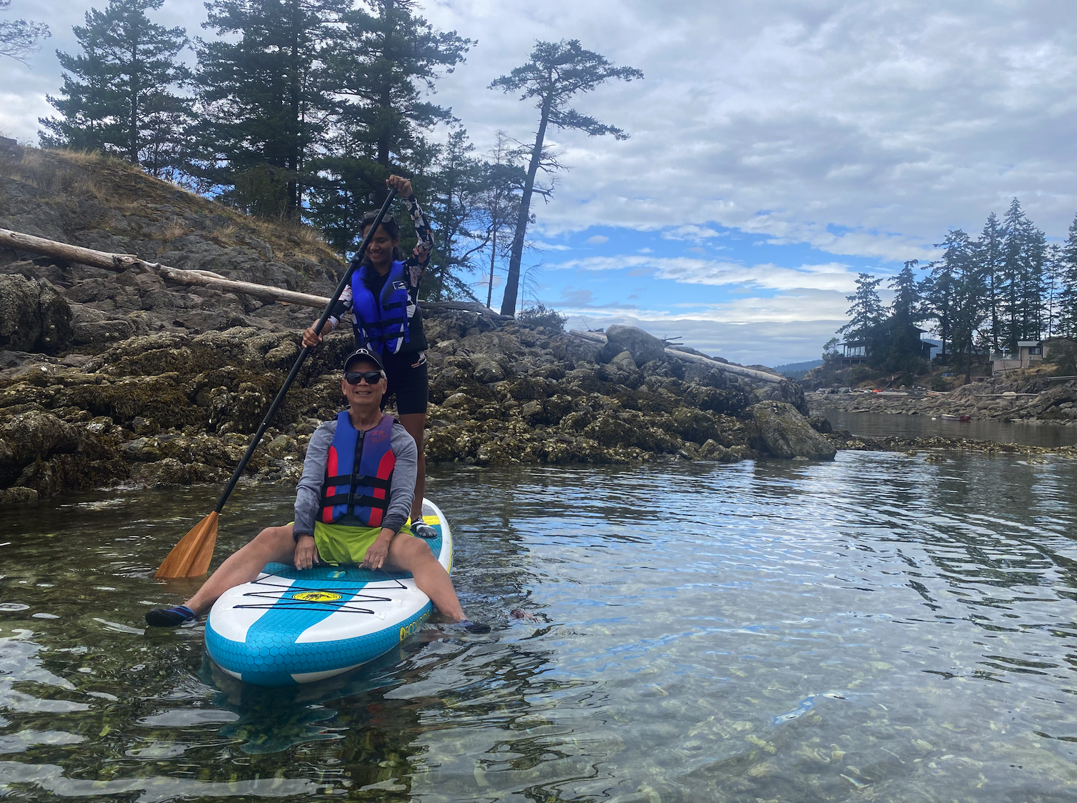Two women are on a paddleboard in a rocky lake, wearing life jackets and smiling. One is standing with a paddle, and the other is sitting at the front of the board.