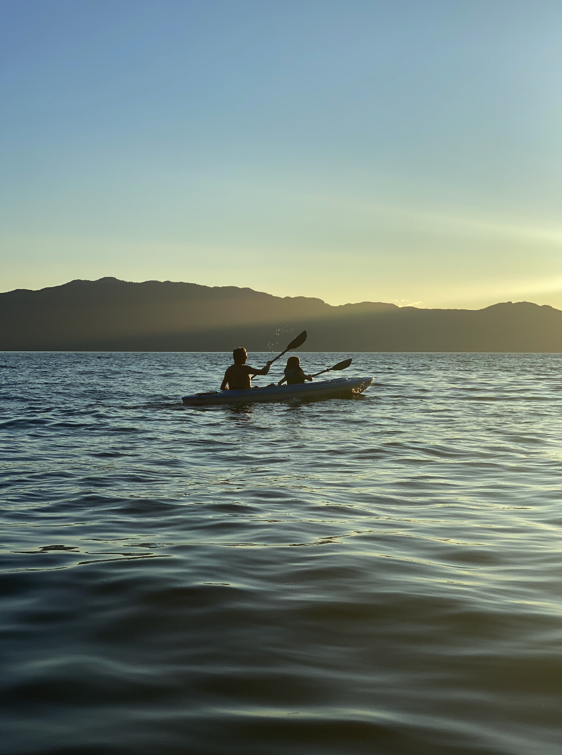 Two people kayaking on a calm body of water during sunset with mountains in the background.