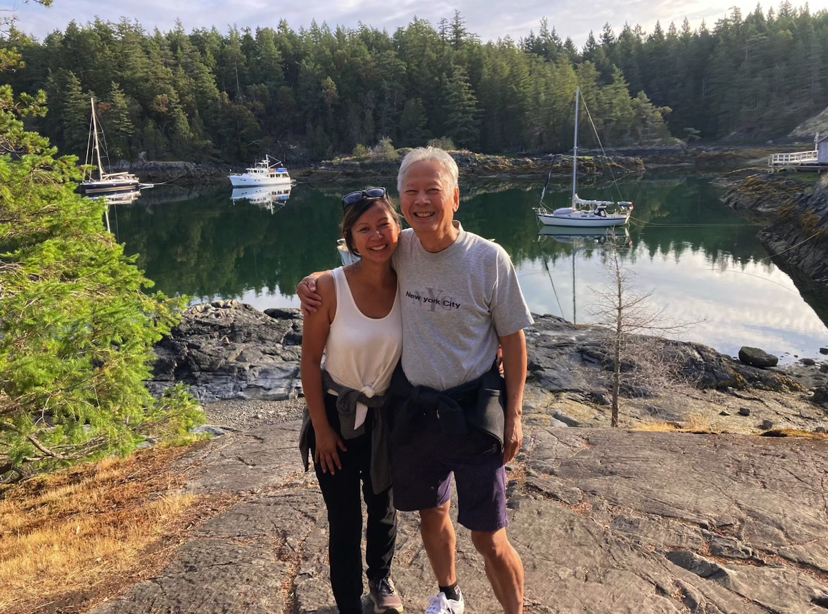 Two smiling people, one young woman and one older man, standing on a rocky shoreline with boats and a forested area reflected in the water behind them.