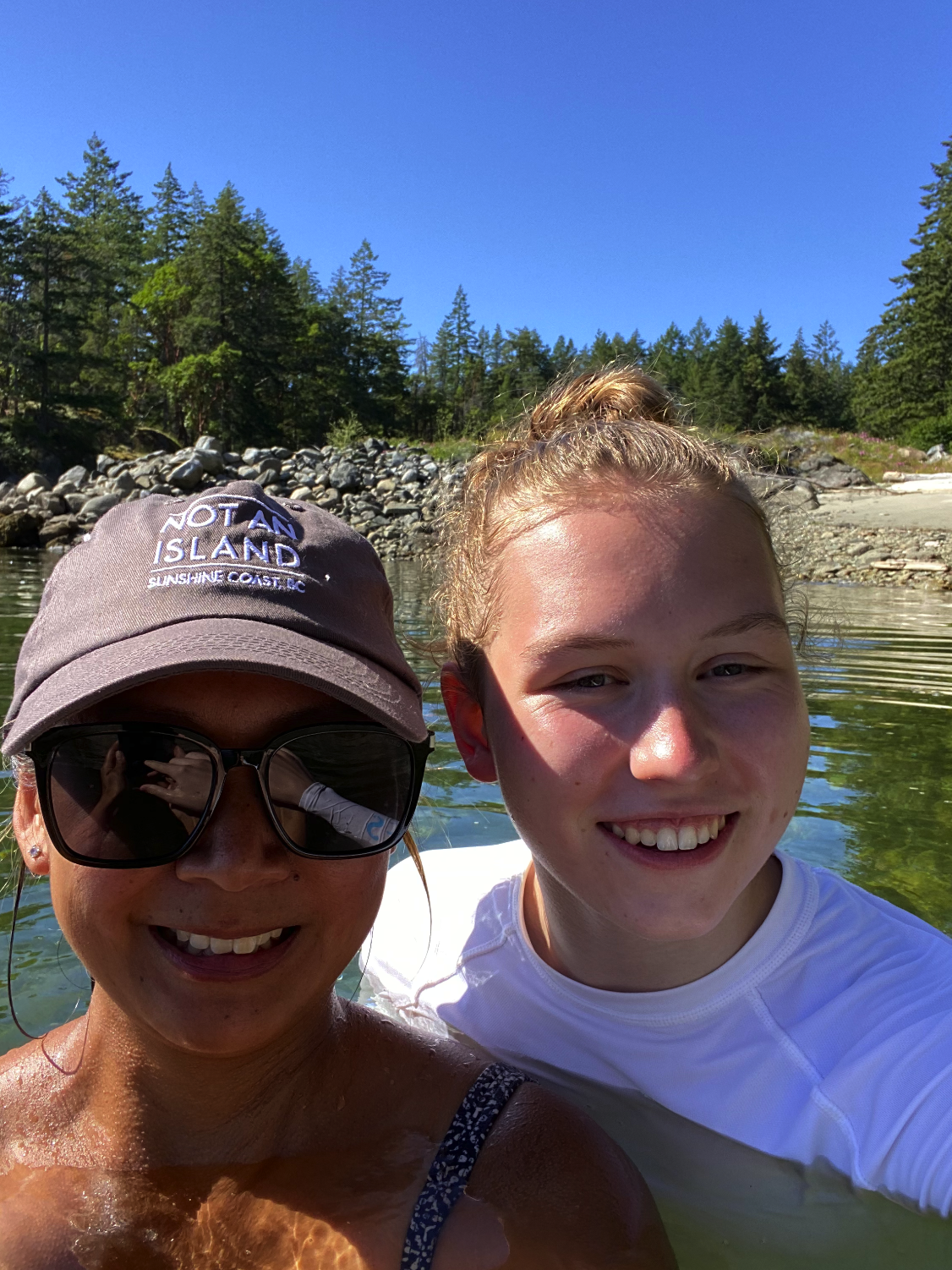 Two women smiling and taking a selfie in a river with a forest and blue sky in the background.
