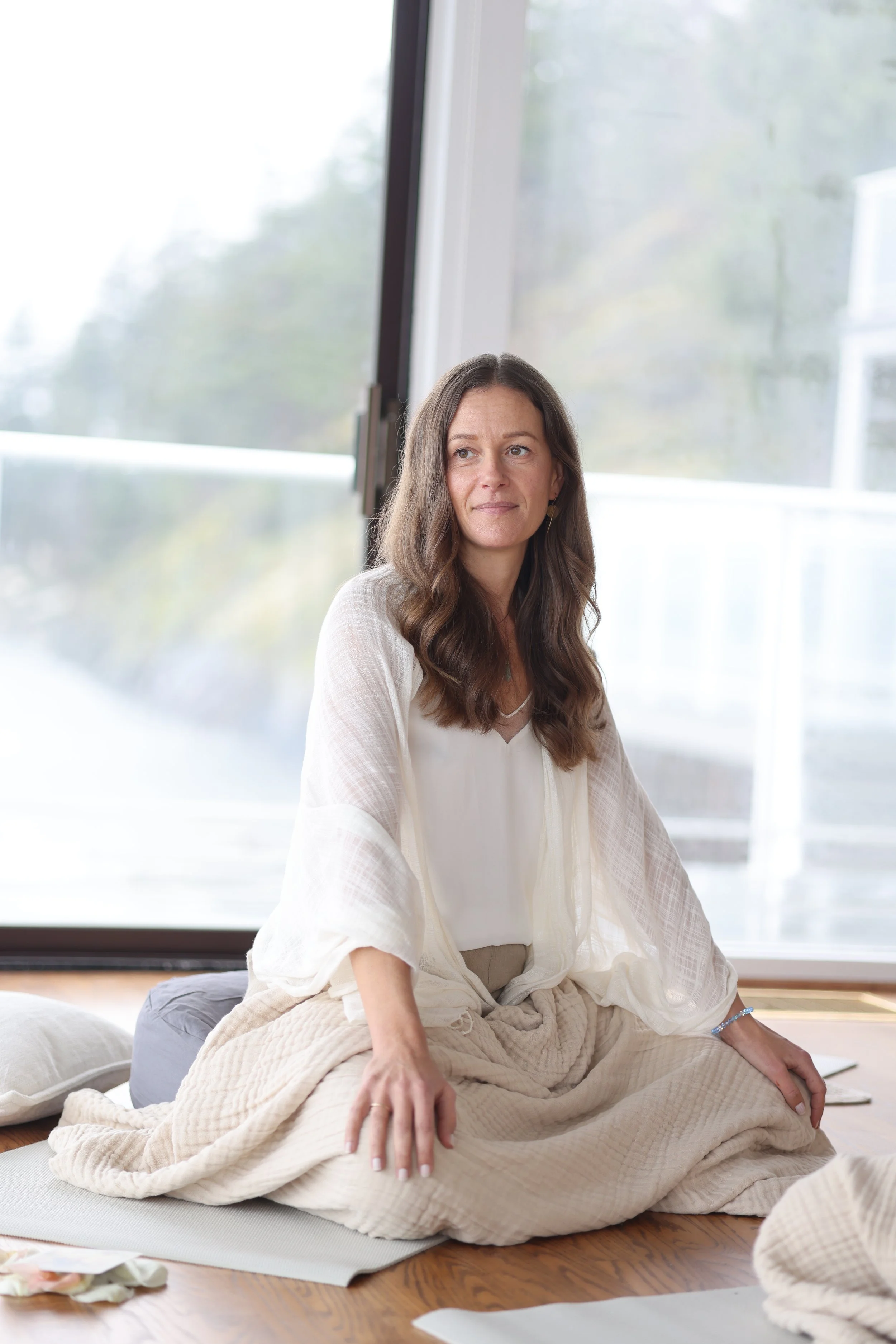 A woman sitting cross-legged on a mat in front of large windows, looking calmly to her side.
