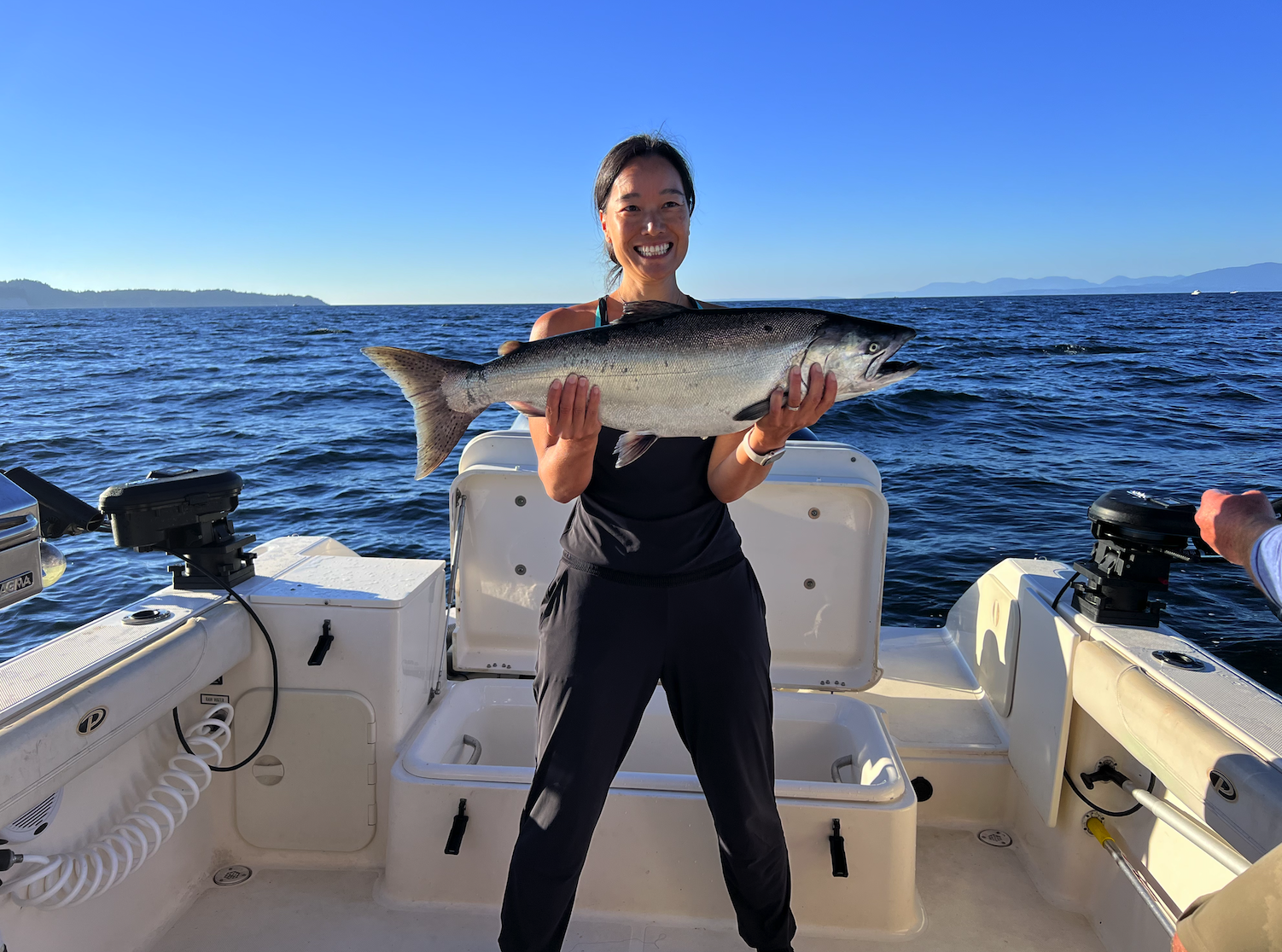 Woman on a boat holding a large fish, smiling with a blue ocean and distant land in the background