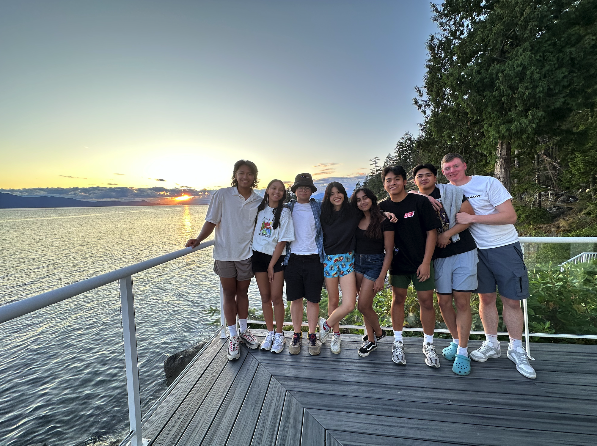 Group of nine friends standing on a dock at sunset with water and trees in the background.
