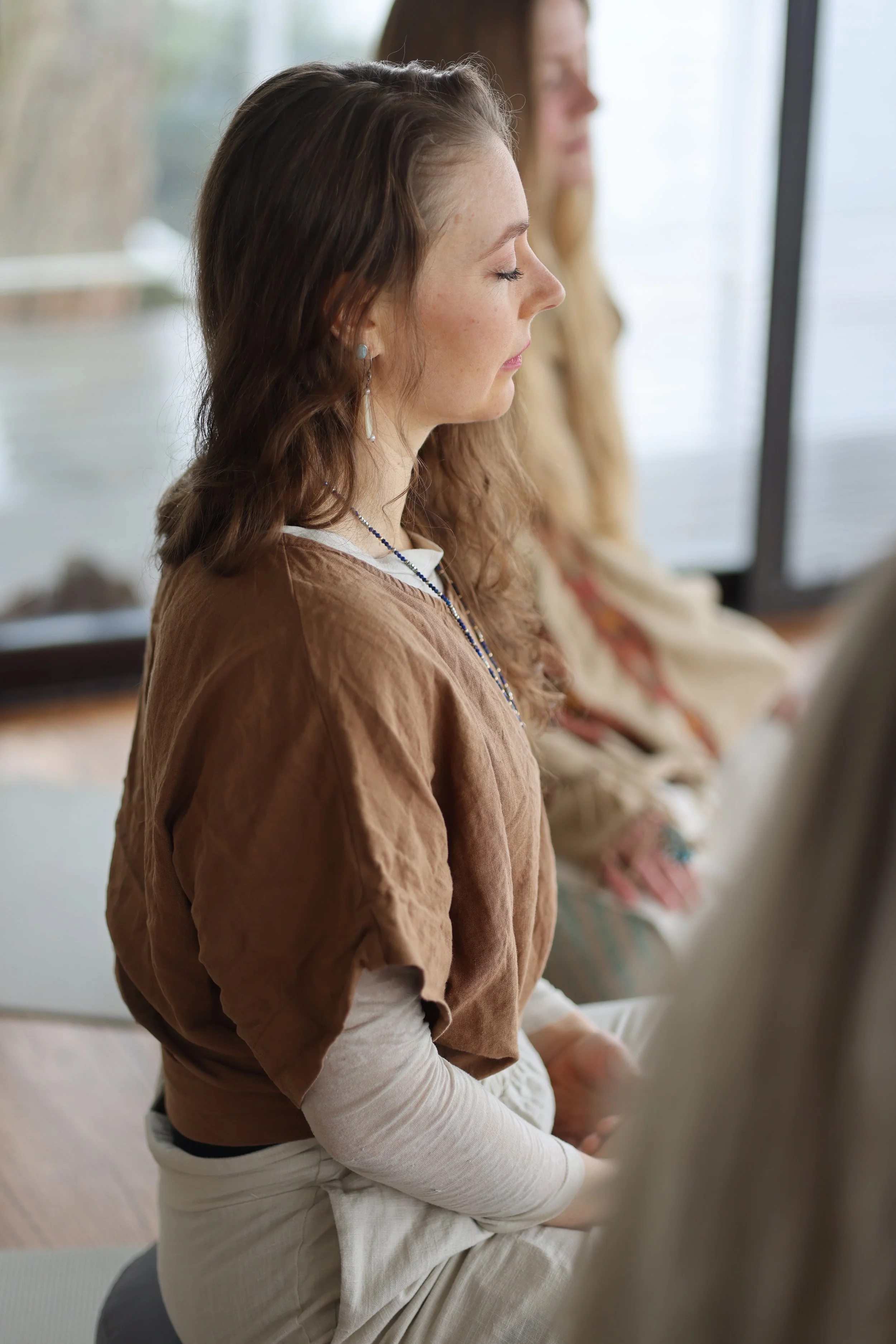 A woman with brown wavy hair, wearing a brown top and beige pants, sitting cross-legged with eyes closed, meditating indoors near large windows with another woman with long blonde hair sitting beside her.