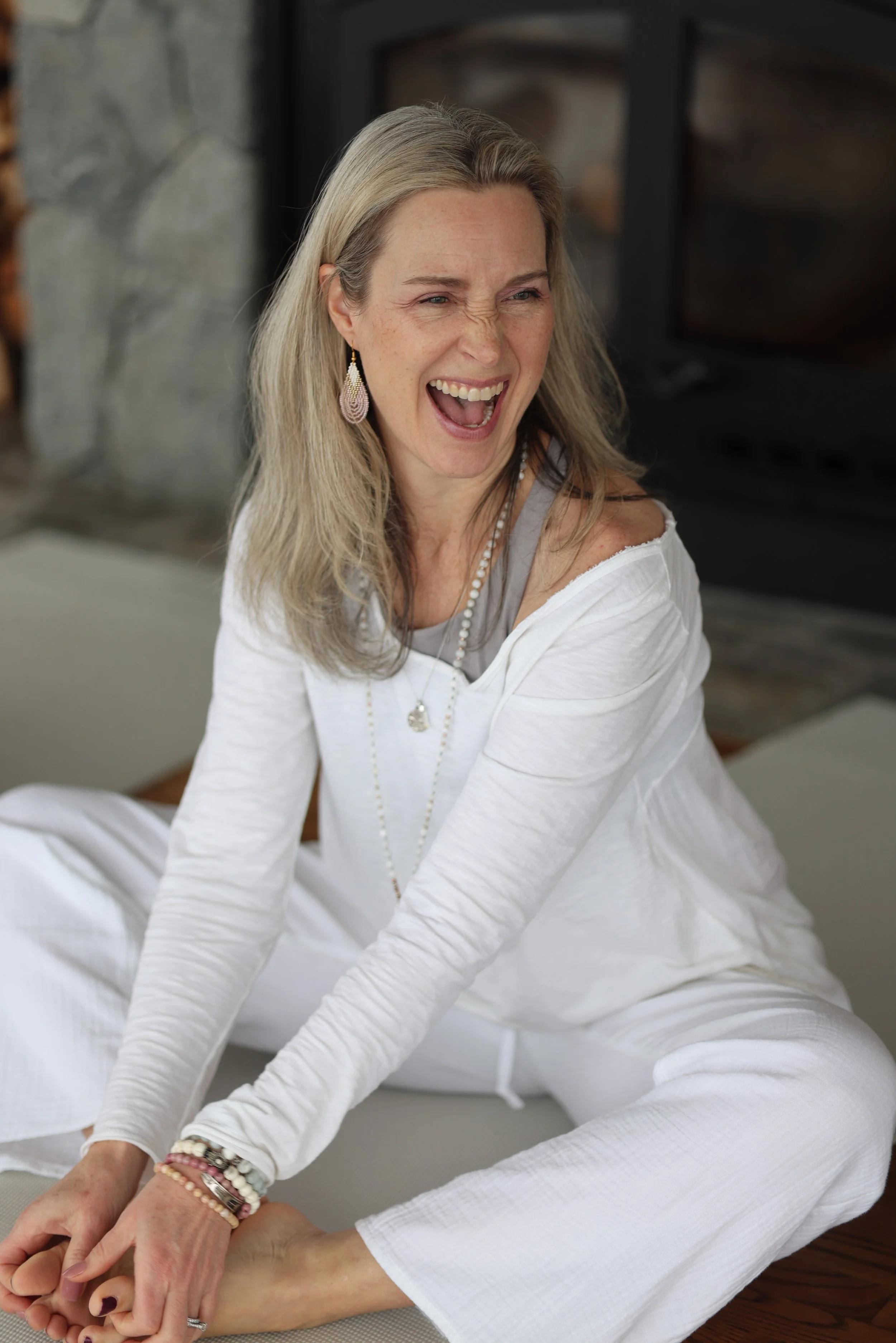 A woman with gray hair, wearing white clothing, sitting cross-legged on the floor, laughing with her mouth open, in front of a fireplace.