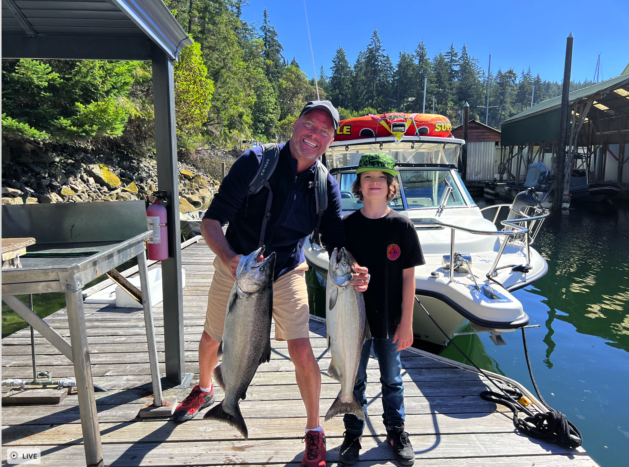 A man and a boy holding caught fish on a dock near a marina, with boats and trees in the background.
