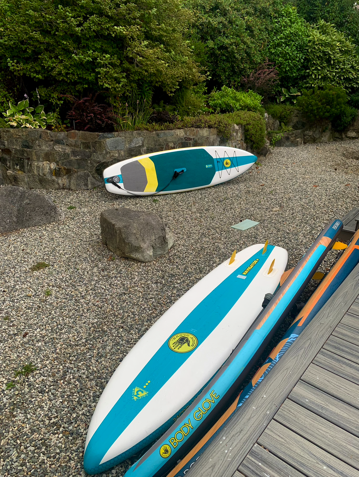 Two stand-up paddleboards resting on gravel next to a wooden deck and a stone wall with lush green bushes in the background.