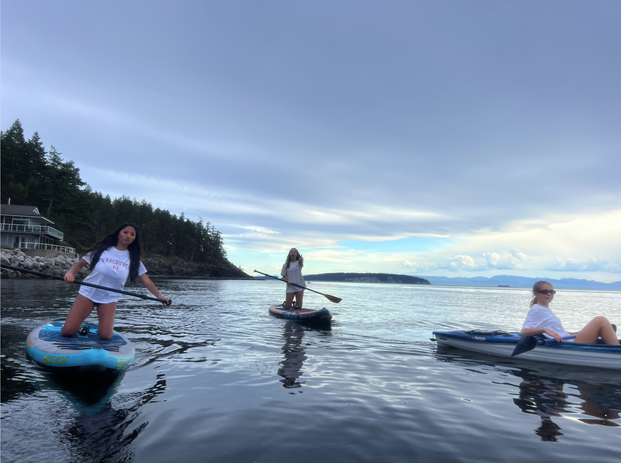 Three women paddleboarding and kayaking on calm water near a shoreline with trees and houses, under a partly cloudy sky.