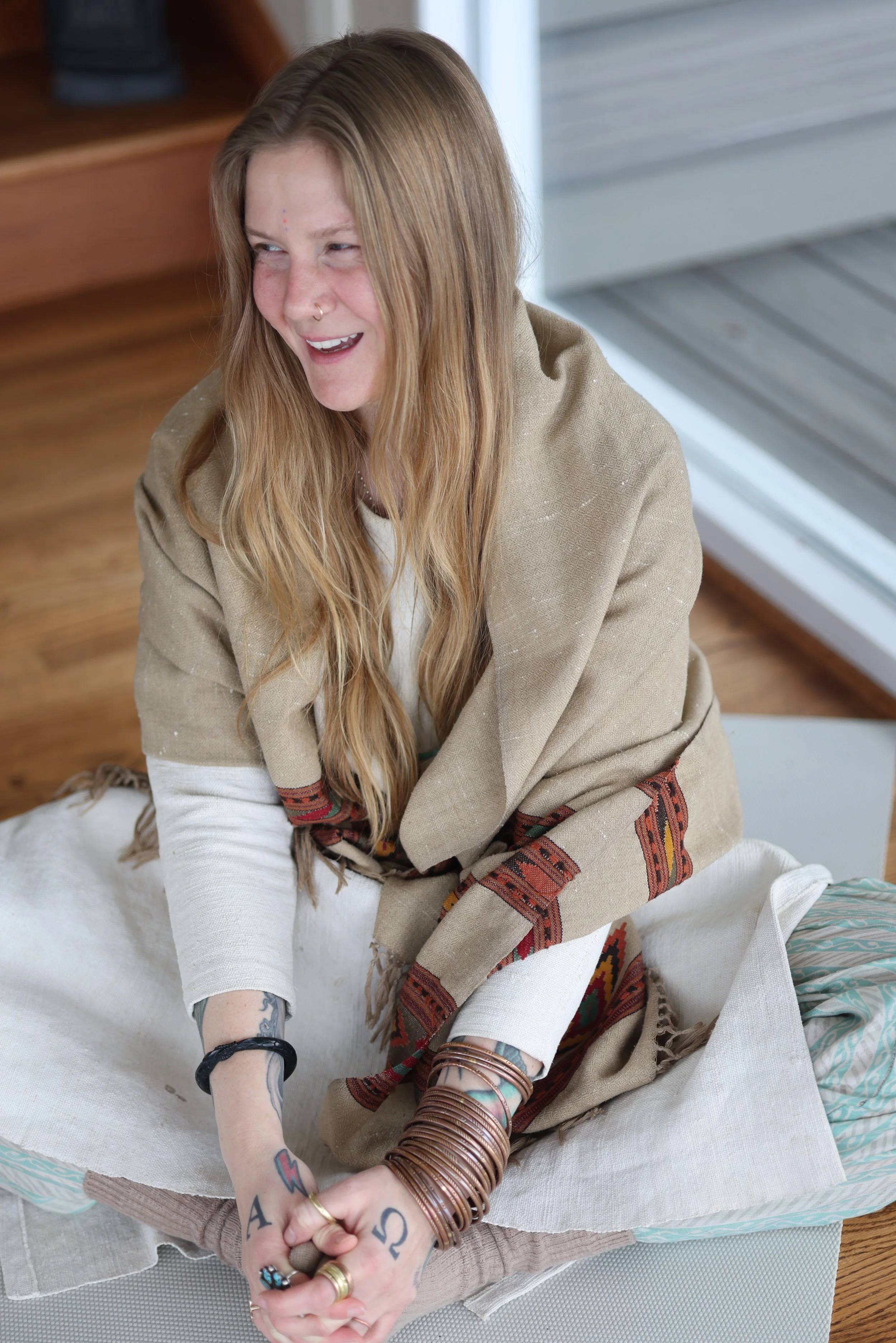 A woman with long red hair, tattoos, and multiple bracelets on her wrists, smiling and sitting on a cloth-covered surface near a window with a wooden floor and furniture nearby.