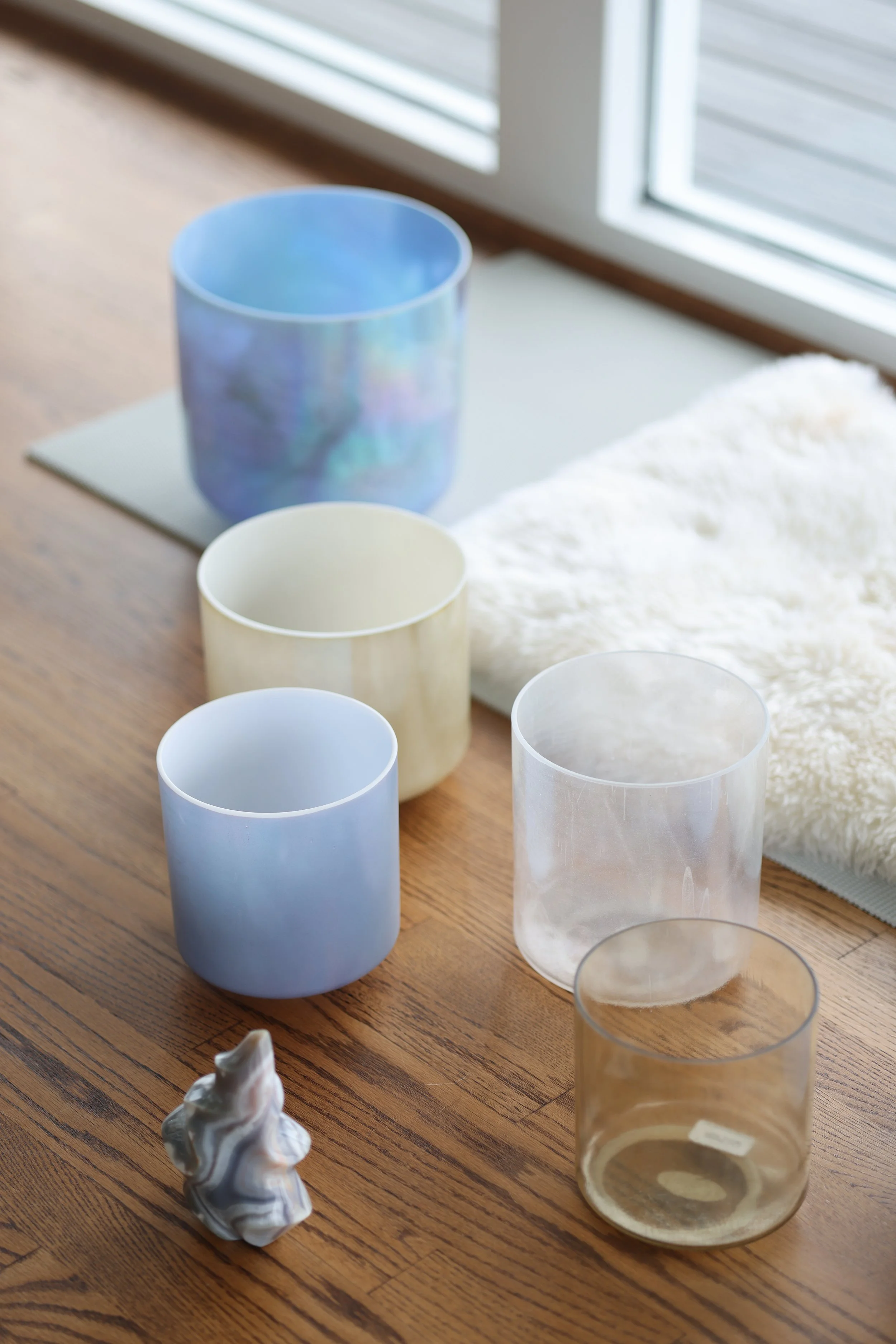 Six colorful glass cups arranged on a wooden table near a window, with a white fluffy cloth and a small decorative item in the foreground.