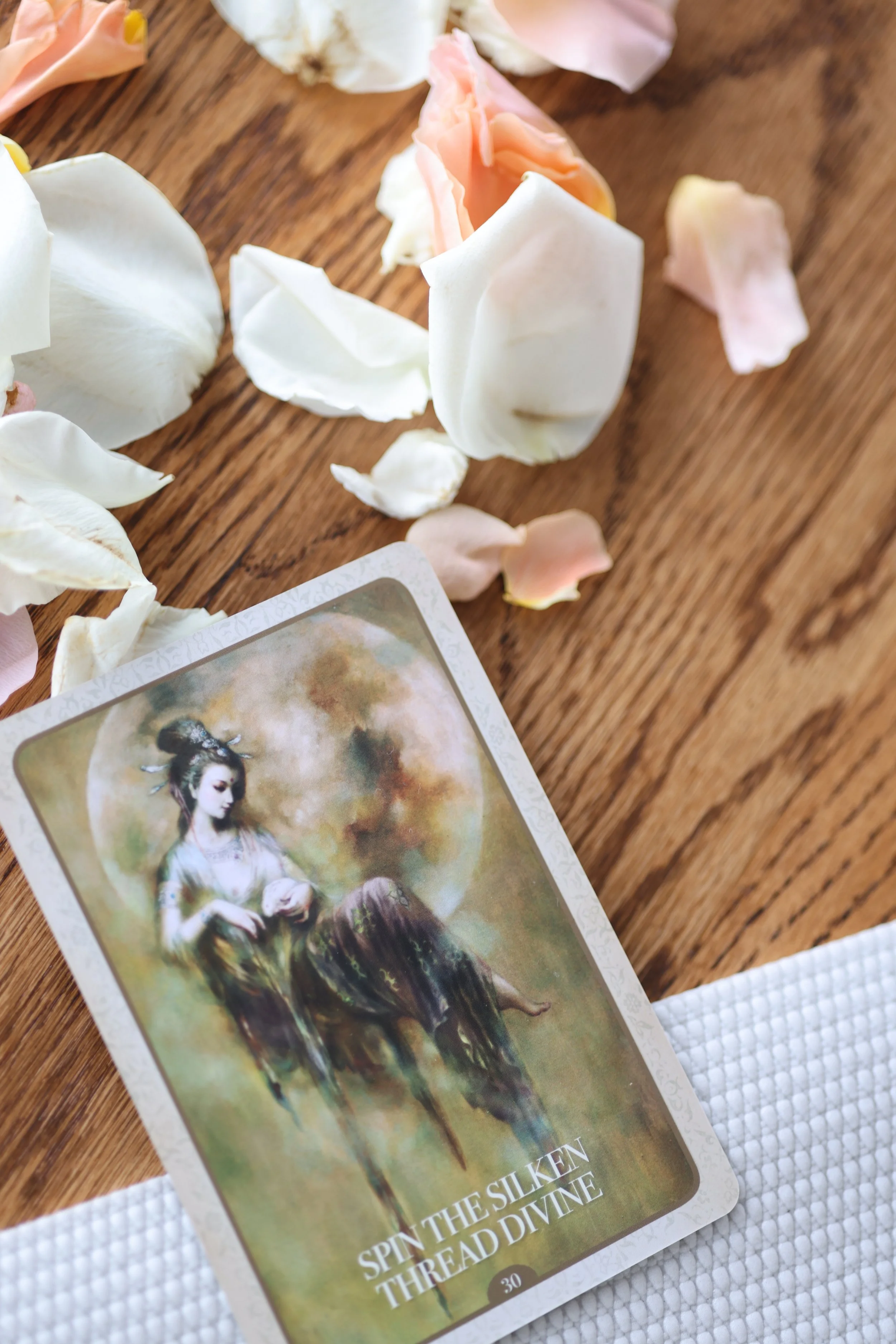 A card titled 'Spin the Silken Thread Divine' resting on a white textured cloth, surrounded by scattered pink and white rose petals on a wooden surface.