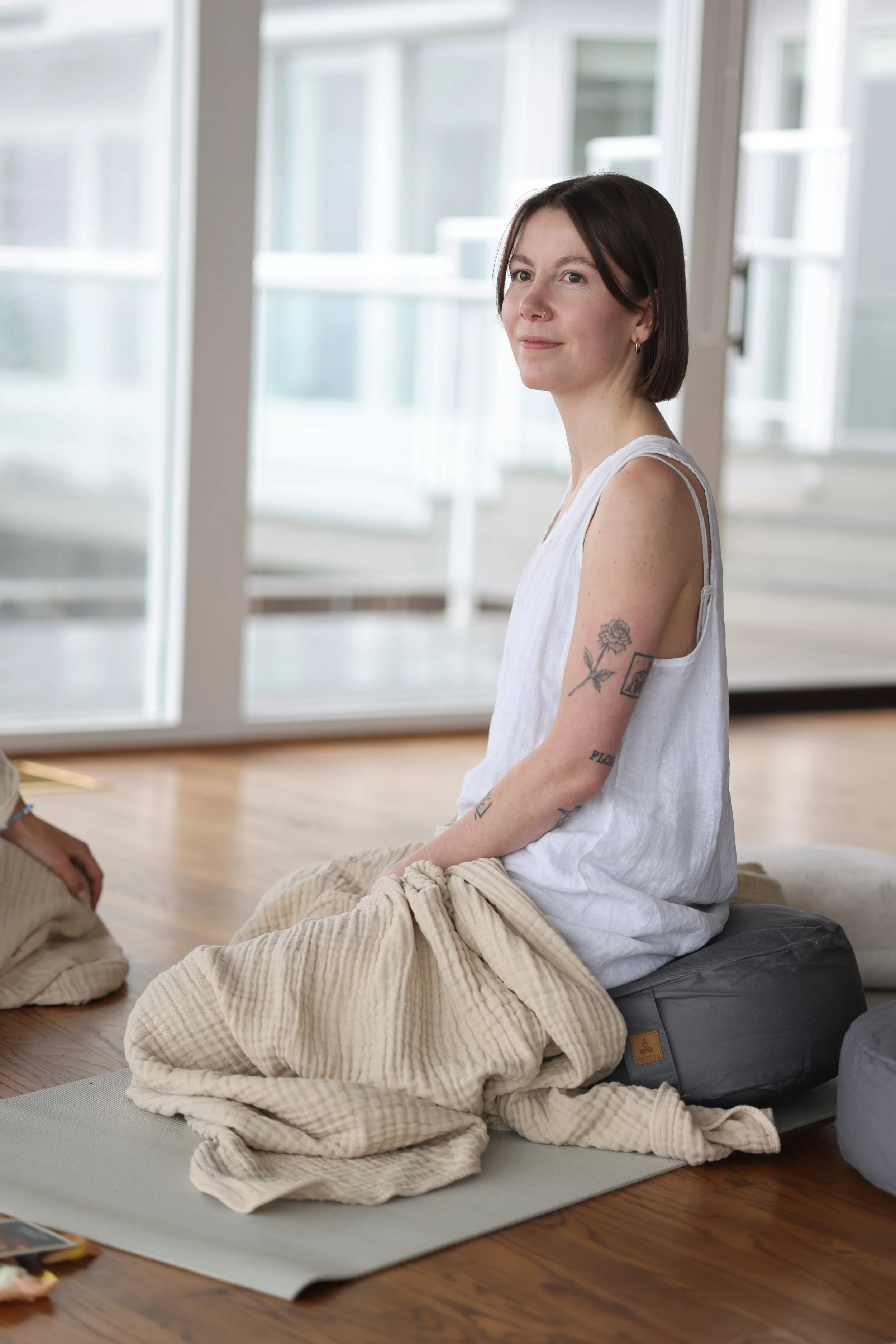 A woman with dark brown hair and tattoos on her arm sits on a cushion on a yoga mat in a bright room with large glass doors, wearing a white sleeveless top and beige pants.
