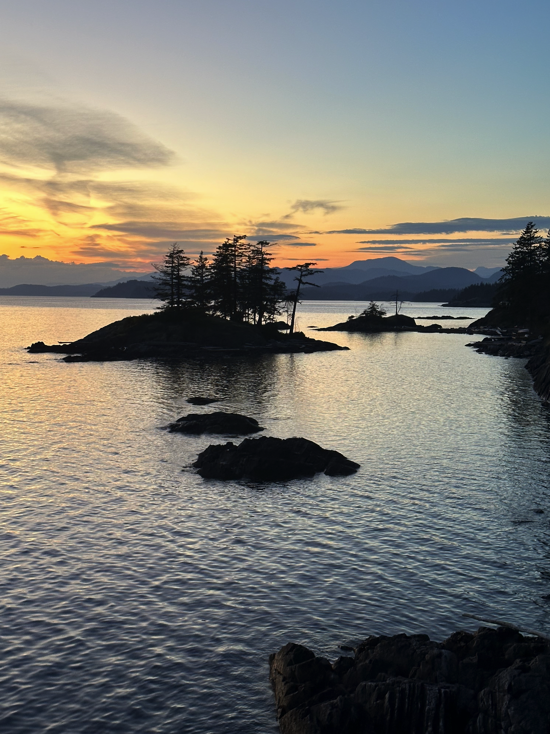 A scenic view of a lake during sunset, with small island and trees silhouetted against the colorful sky and distant mountains.
