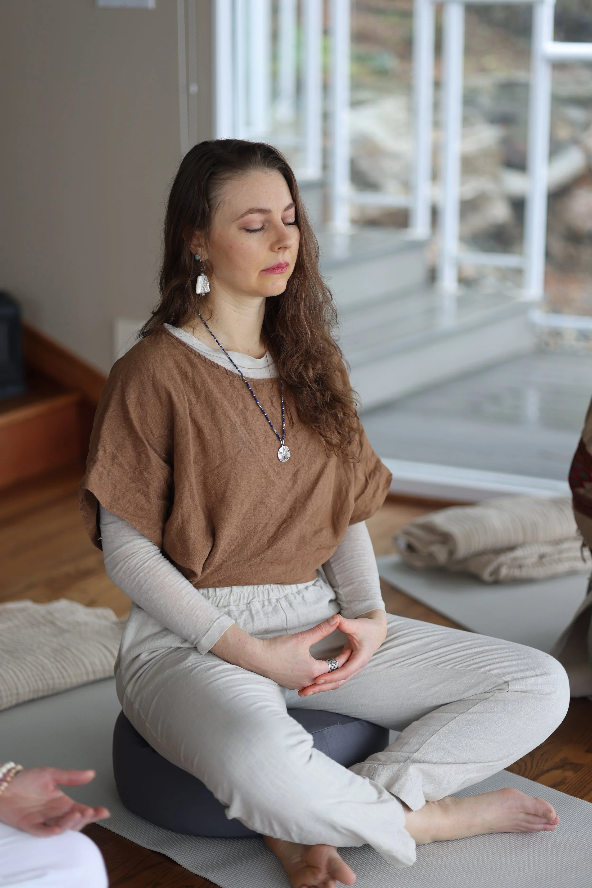 A woman with long brown hair, sitting cross-legged on a yoga mat, meditating with her eyes closed indoors near large windows.