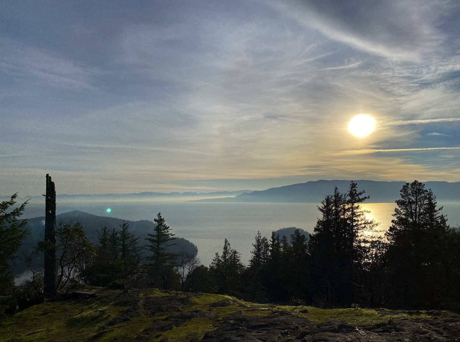 Sunrise over mountains and a body of water, with trees in the foreground and a clear sky with some clouds.