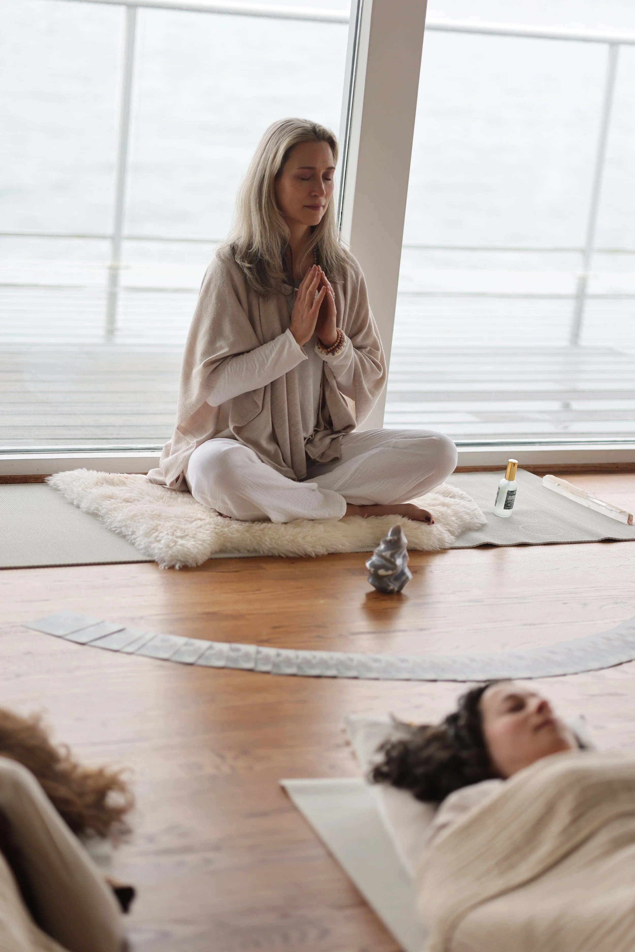 A woman practicing yoga in a serene space by a large window, with her eyes closed and hands in a prayer position, sitting on a mat with a fur blanket, while a person lies on a mat nearby, and spiritual items like a statue and essential oil bottles ar