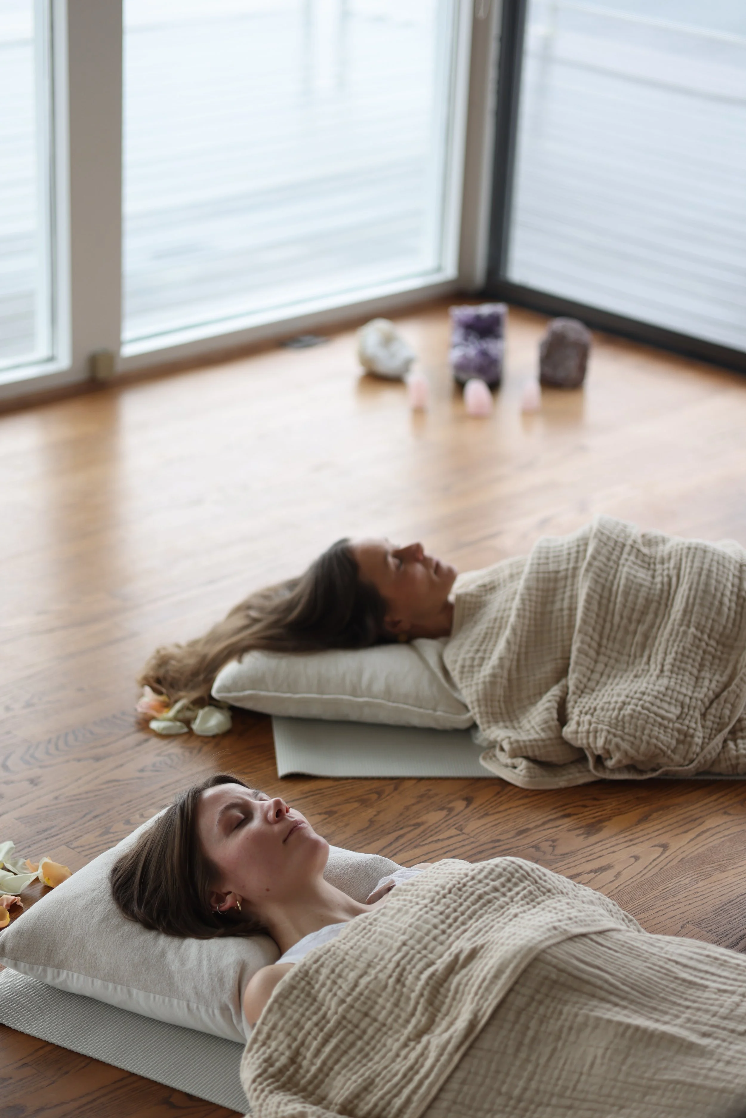 Two women are lying on yoga mats on a wooden floor, appearing to meditate with their eyes closed. One woman has long brown hair and the other has short brown hair. Behind them, near a large window, there are various crystals and candles arranged on t