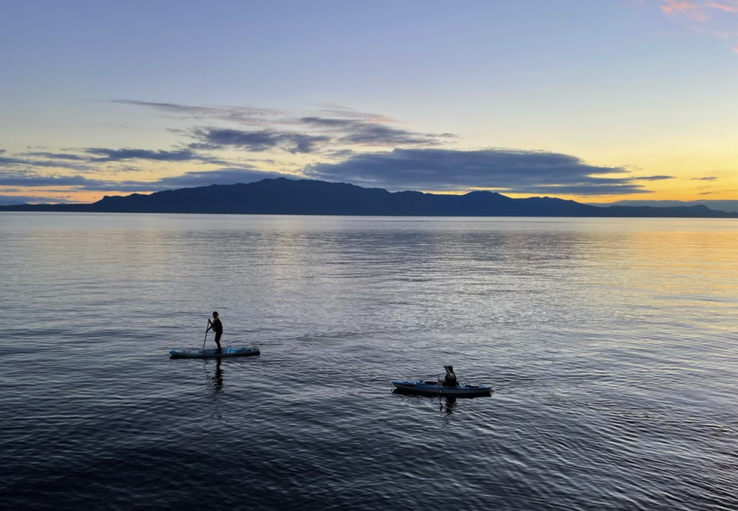 A person paddleboarding and a dog sitting on a kayak on a calm lake during sunset, with mountains in the distance and a partly cloudy sky.