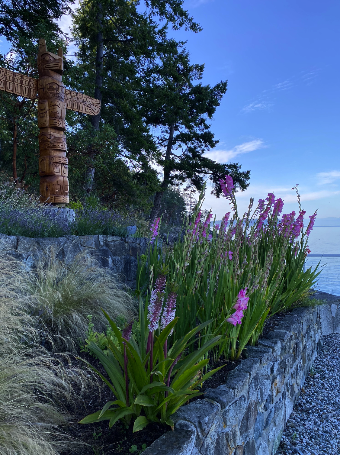 A garden with pink flowers along a stone border, a wooden totem pole, trees, and a body of water in the background under a blue sky.