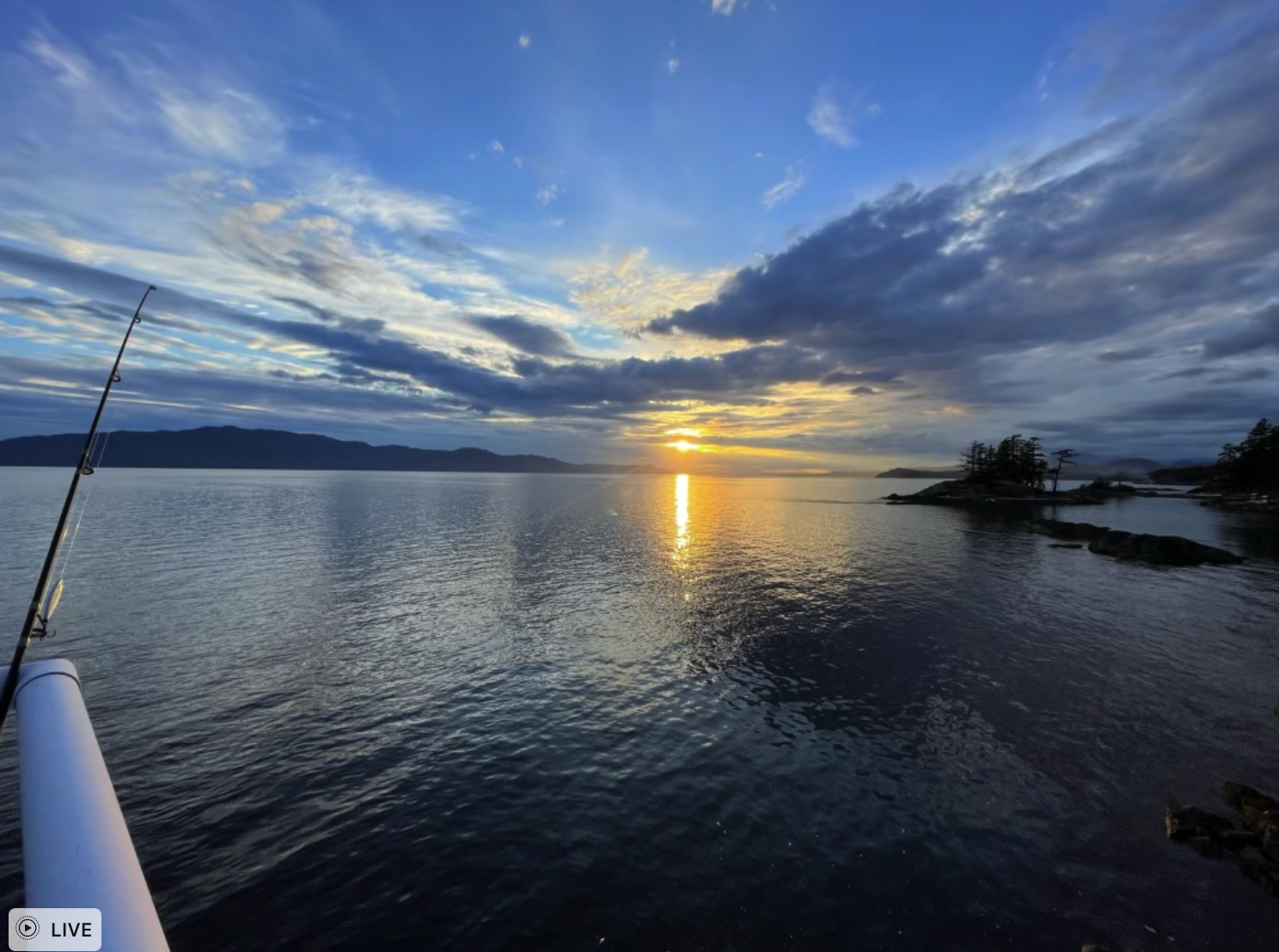 Sunset over a body of water with clouds in the sky and a glimpse of land on the horizon; a fishing rod is visible in the left foreground.