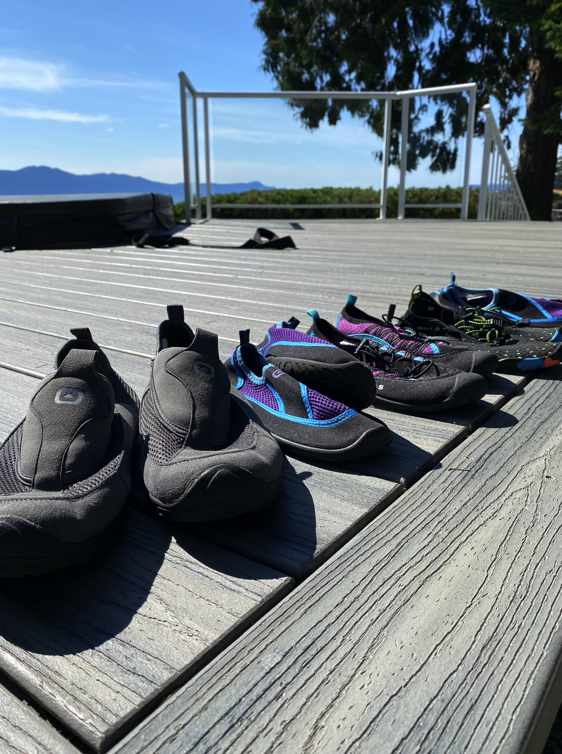 A row of five pairs of climbing shoes arranged on a wooden deck, with mountains and a blue sky in the background.