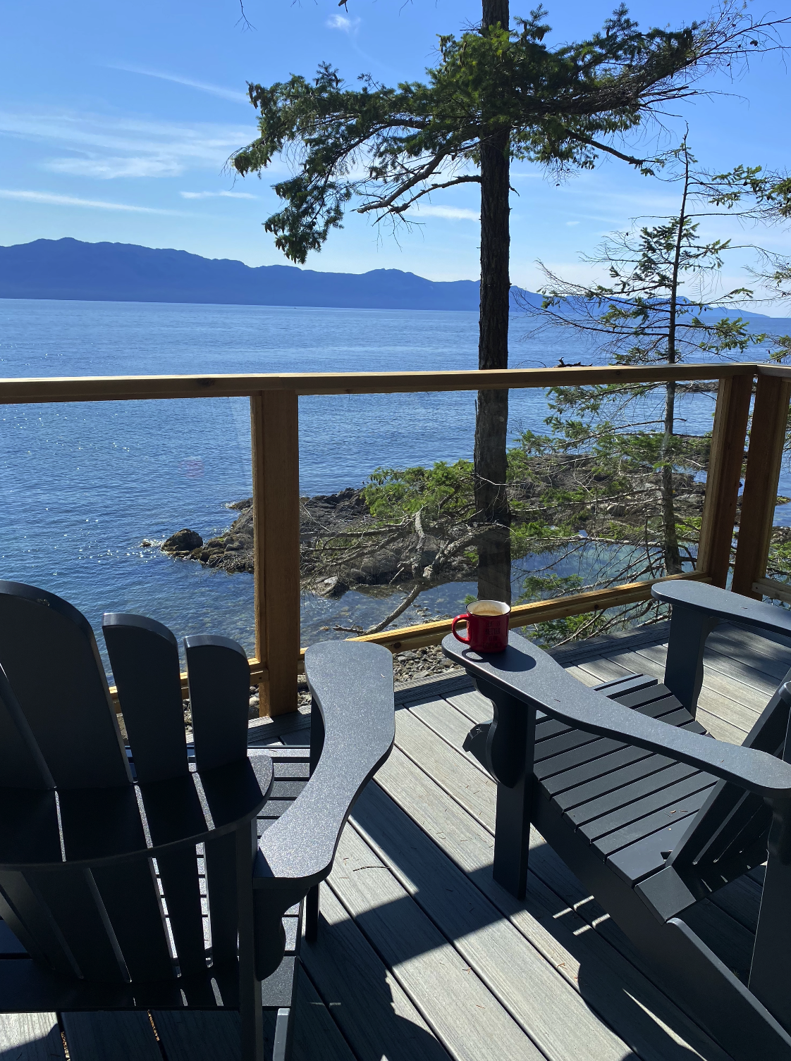 View of a wooden deck overlooking a large body of water with distant mountains and clear blue sky. There are Adirondack chairs and a red mug on the deck.