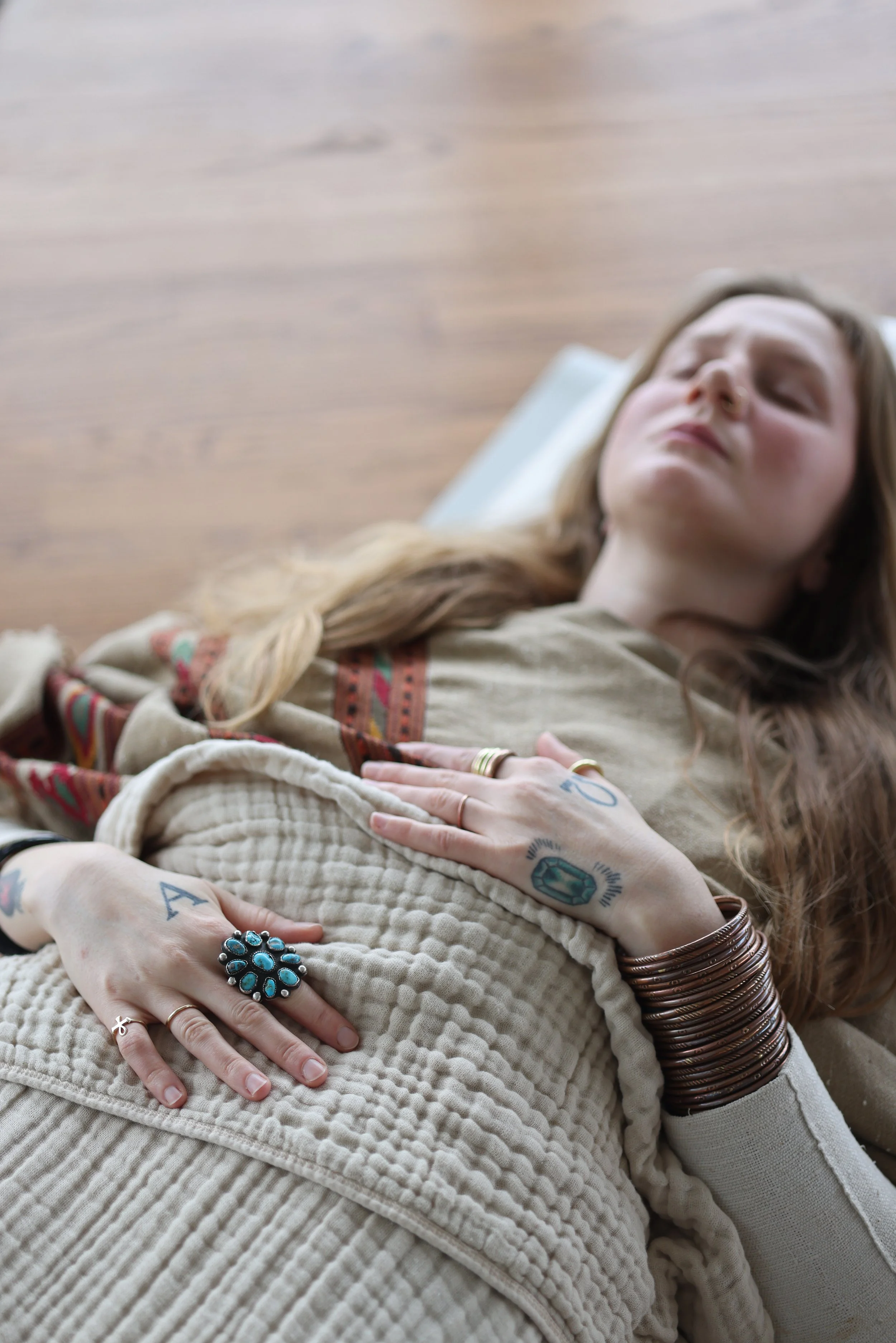 A woman lying on her back with eyes closed, resting on a pillow on a wooden floor, wearing a beige outfit with colorful embroidery, multiple tattoos on her hands, finger rings, and a large turquoise ring, with a stack of copper bangles on her wrist.