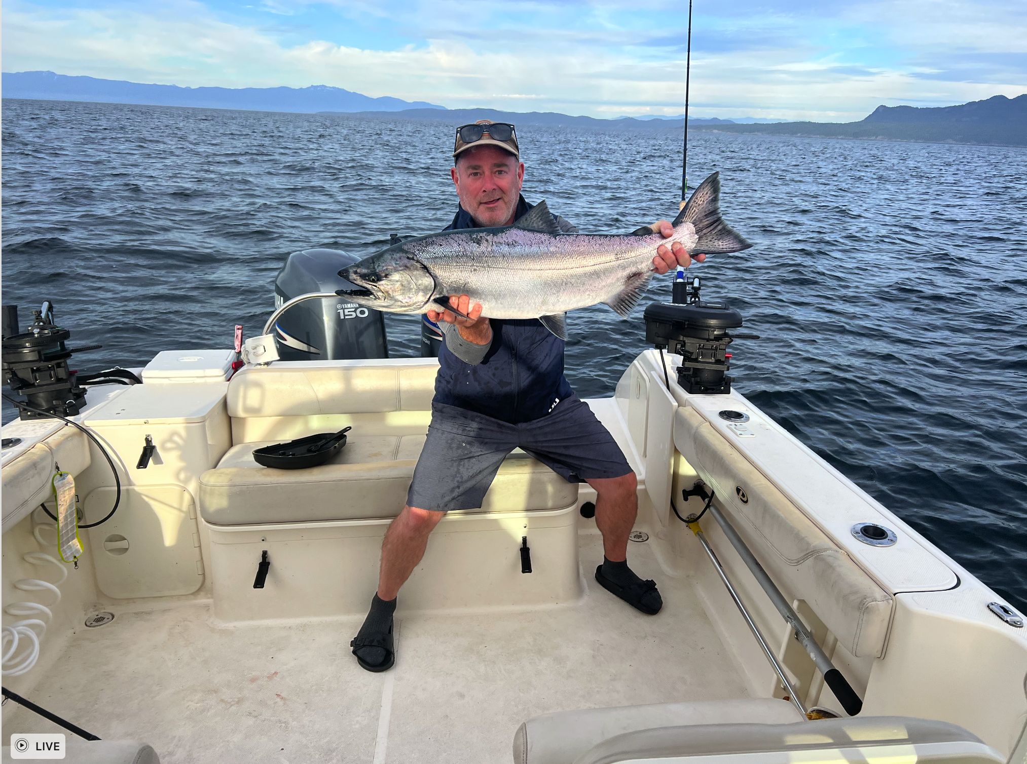 A man on a boat holding a large fish over the water with mountains in the background.