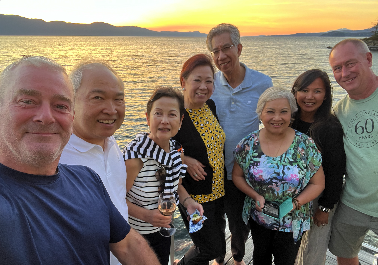 Group of people smiling for a photo on a sunset by a body of water, with mountains in the background.