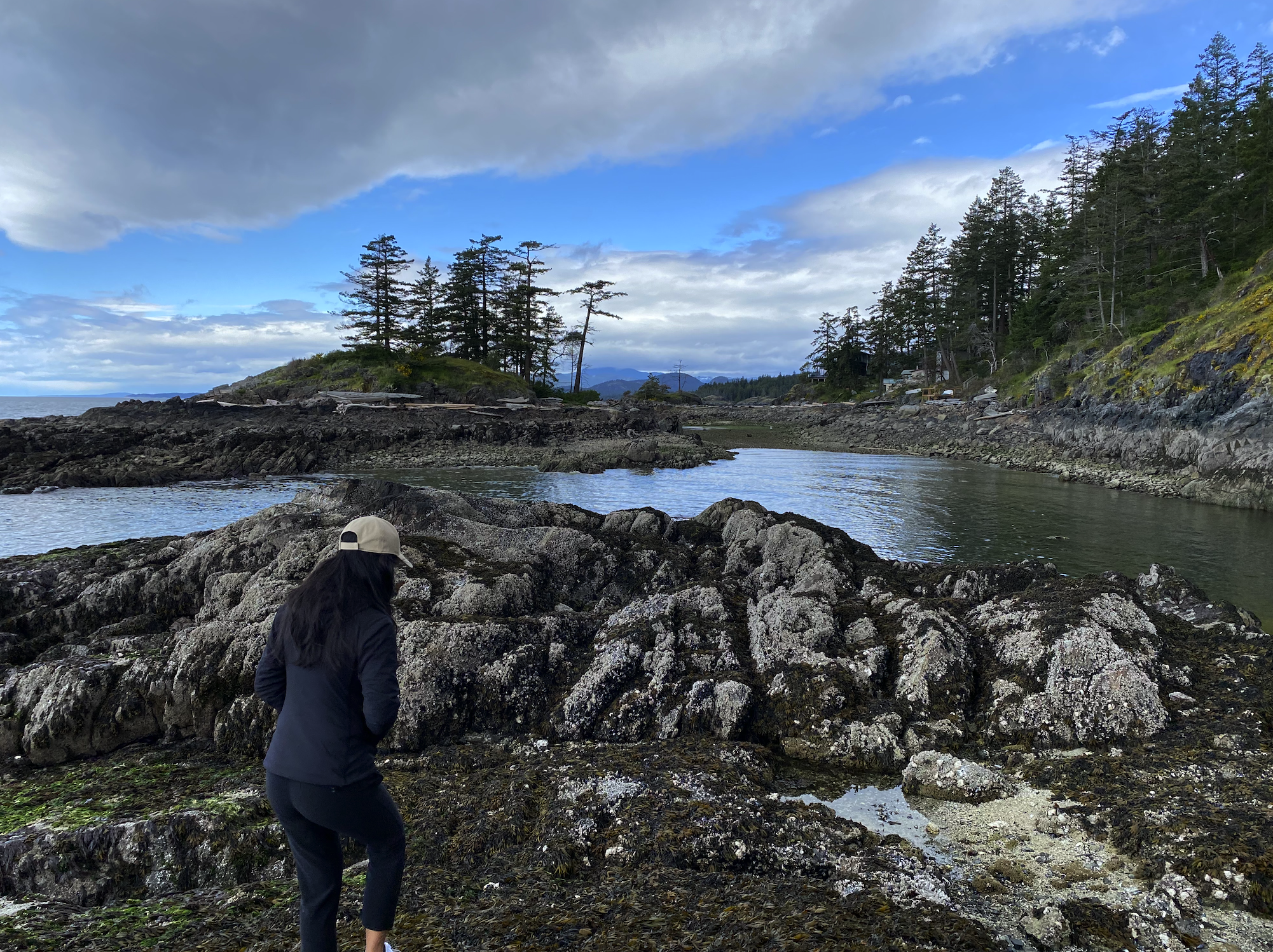 A person with long dark hair, wearing a light-colored cap and dark outdoor clothing, walking on a rocky shoreline near a river, with forested hills and a partly cloudy sky in the background.