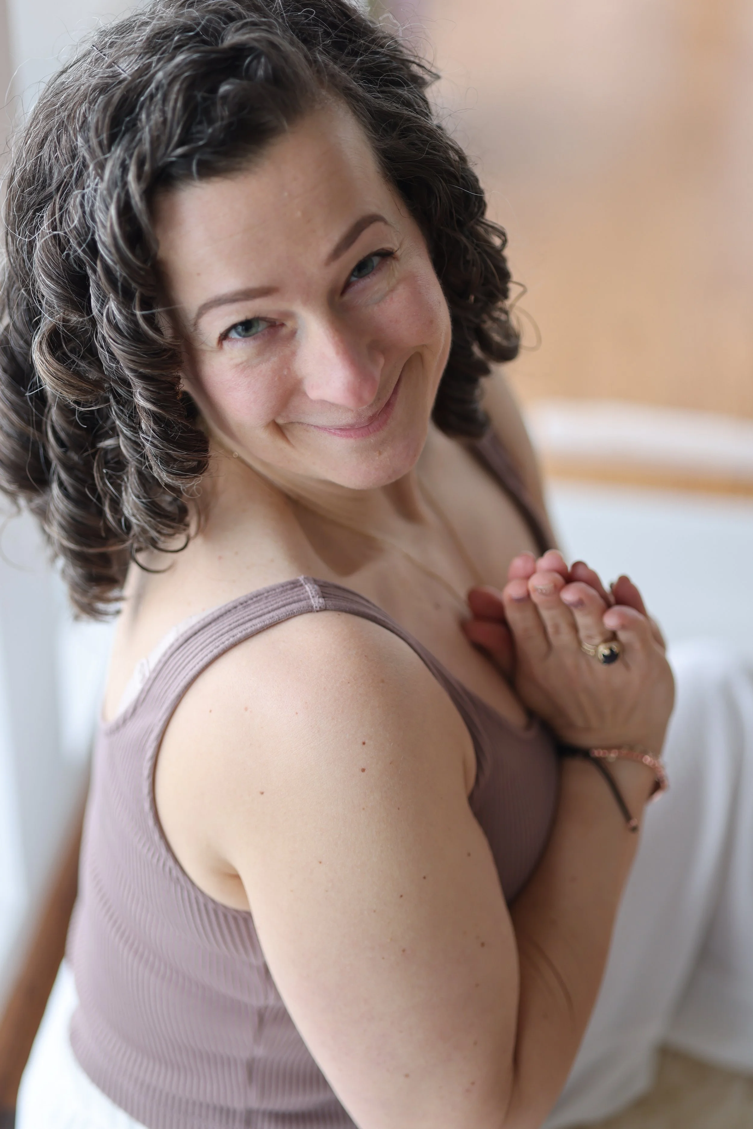A woman with curly brown hair smiling warmly, wearing a sleeveless beige top, holding her hands together near her chest, with a blurred background.