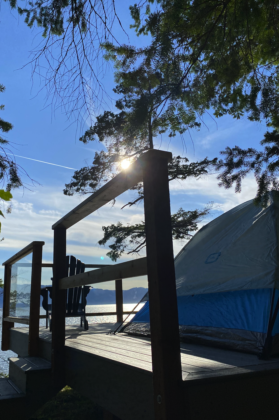Sun shining over a wooden deck with a camping tent and outdoor chair, surrounded by trees and overlooking a body of water with mountains in the distance.