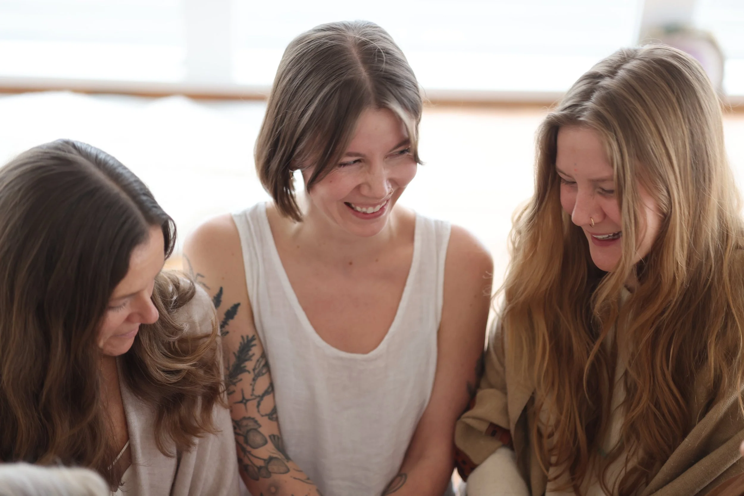 Three women sitting together, laughing and smiling in a bright room.