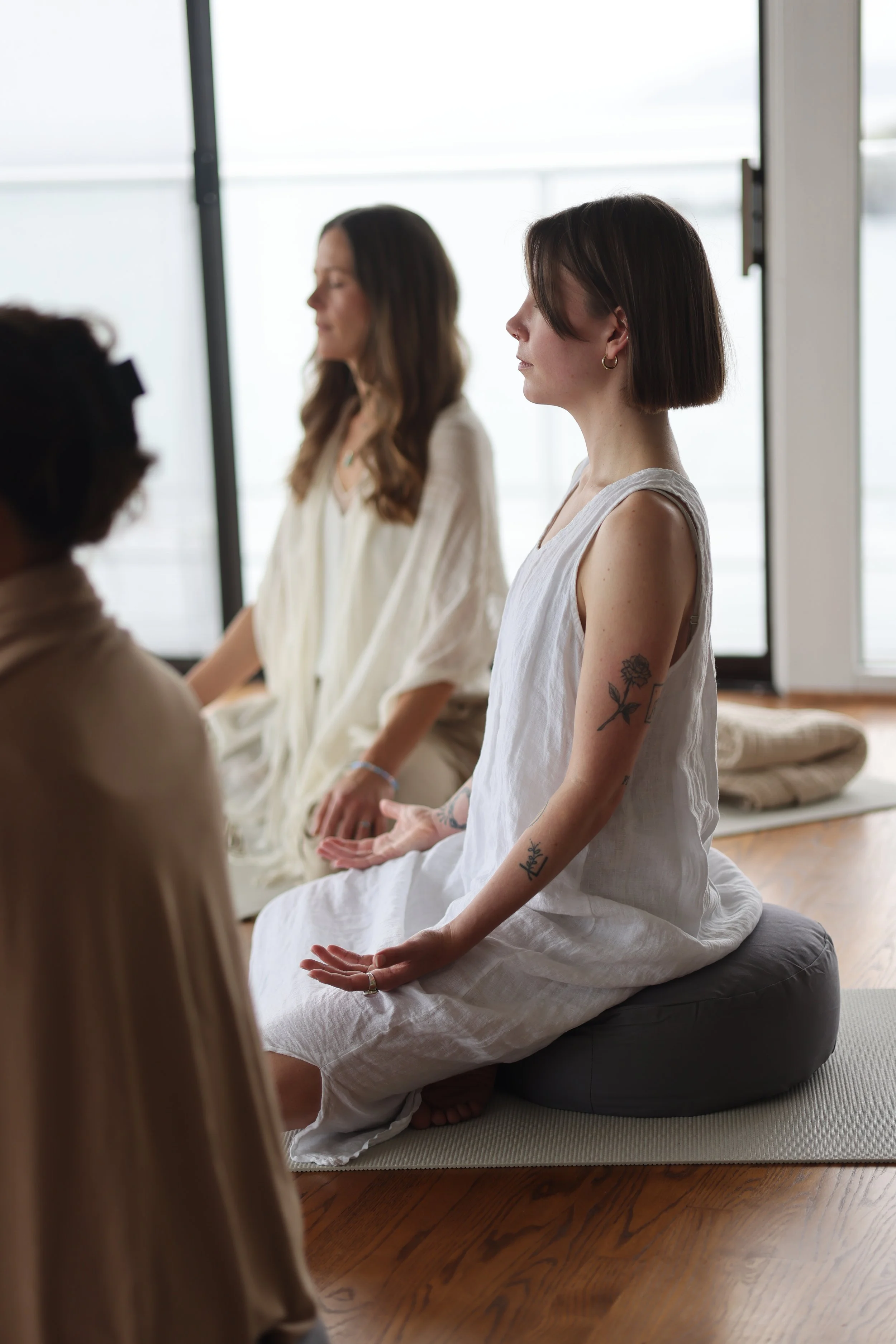 Three women sitting in a meditation pose on mats indoors, with large windows in the background letting in natural light.