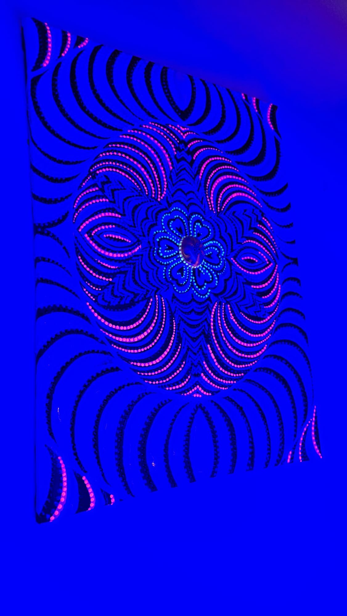 “Colorful symmetrical abstract painting with hypnotic circular patterns and central gem, exploring vibration and energy by artist Laura Garcia.”Colors on it: White, black,pink,blue glow in the dark