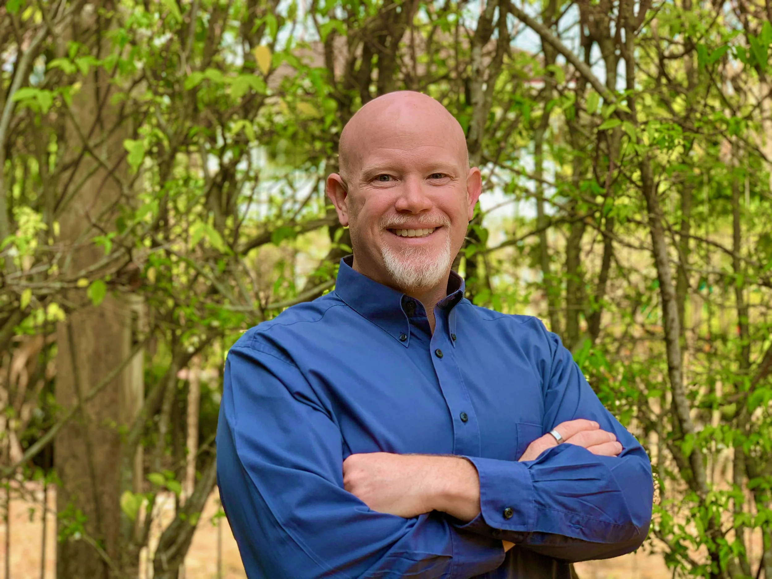 A man with a bald head and goatee smiles with arms crossed, wearing a blue button-down shirt, outdoors in front of green foliage.