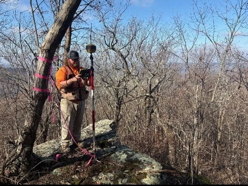 A researcher standing on a rocky hilltop with a surveying device, surrounded by leafless trees, conducting a field study.