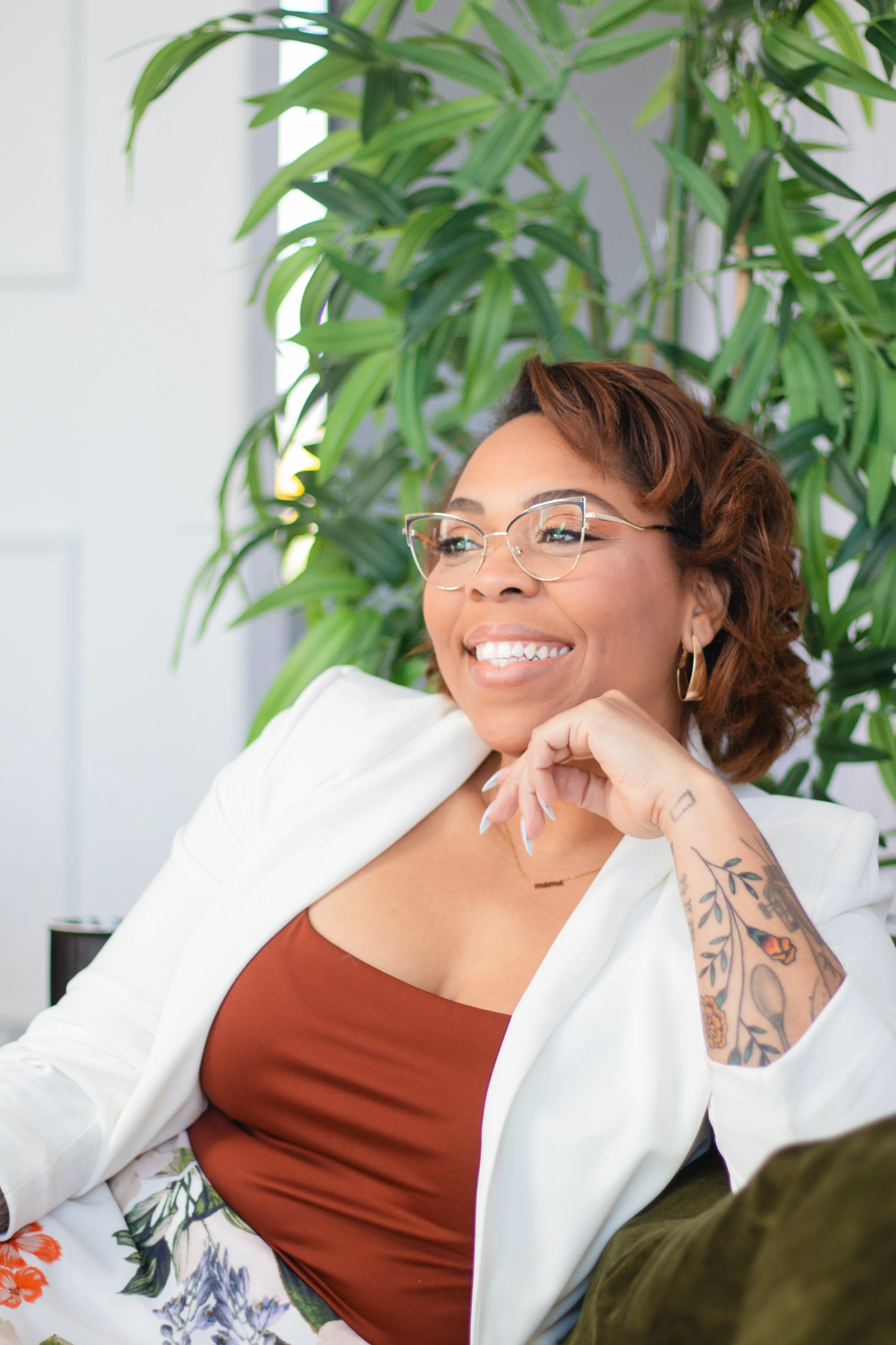 Woman with short auburn hair, glasses, and tattoos on her arm, smiling while sitting in front of a large green plant indoors.