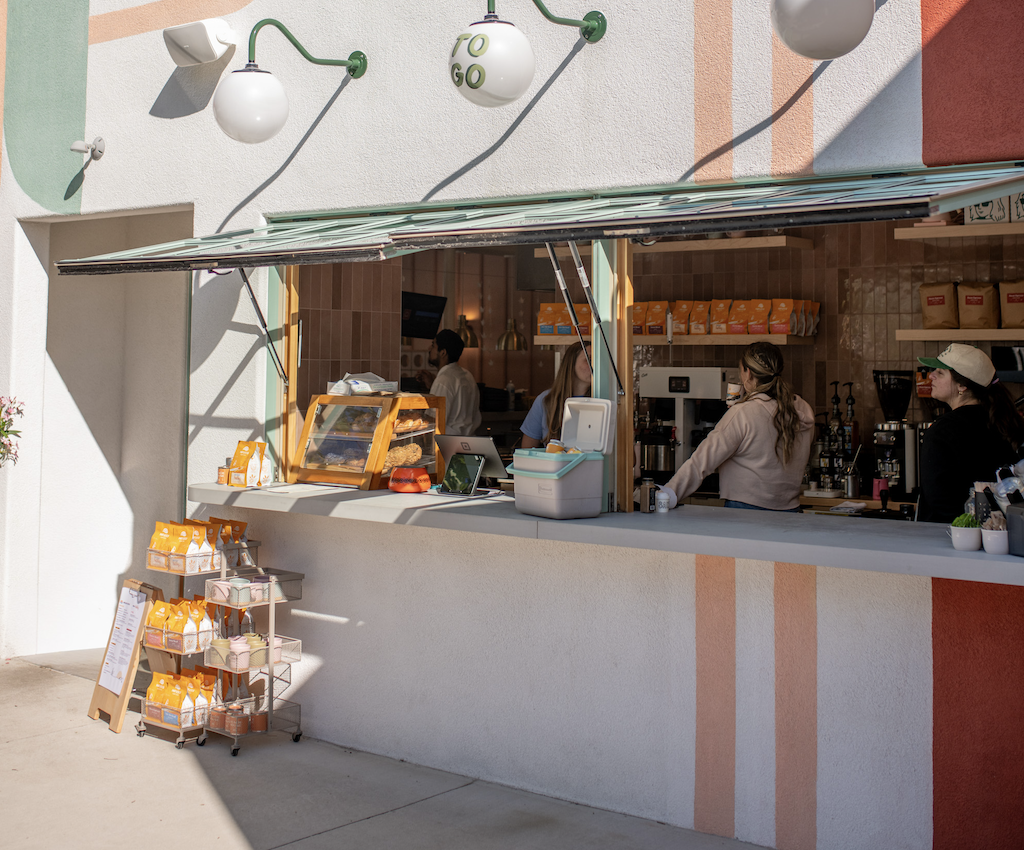 A coffee stand with an open window, showing baristas inside preparing drinks. Outside, a display with bags of coffee and tea, and a menu board. The building's exterior is decorated with pastel-colored stripes and modern lighting.