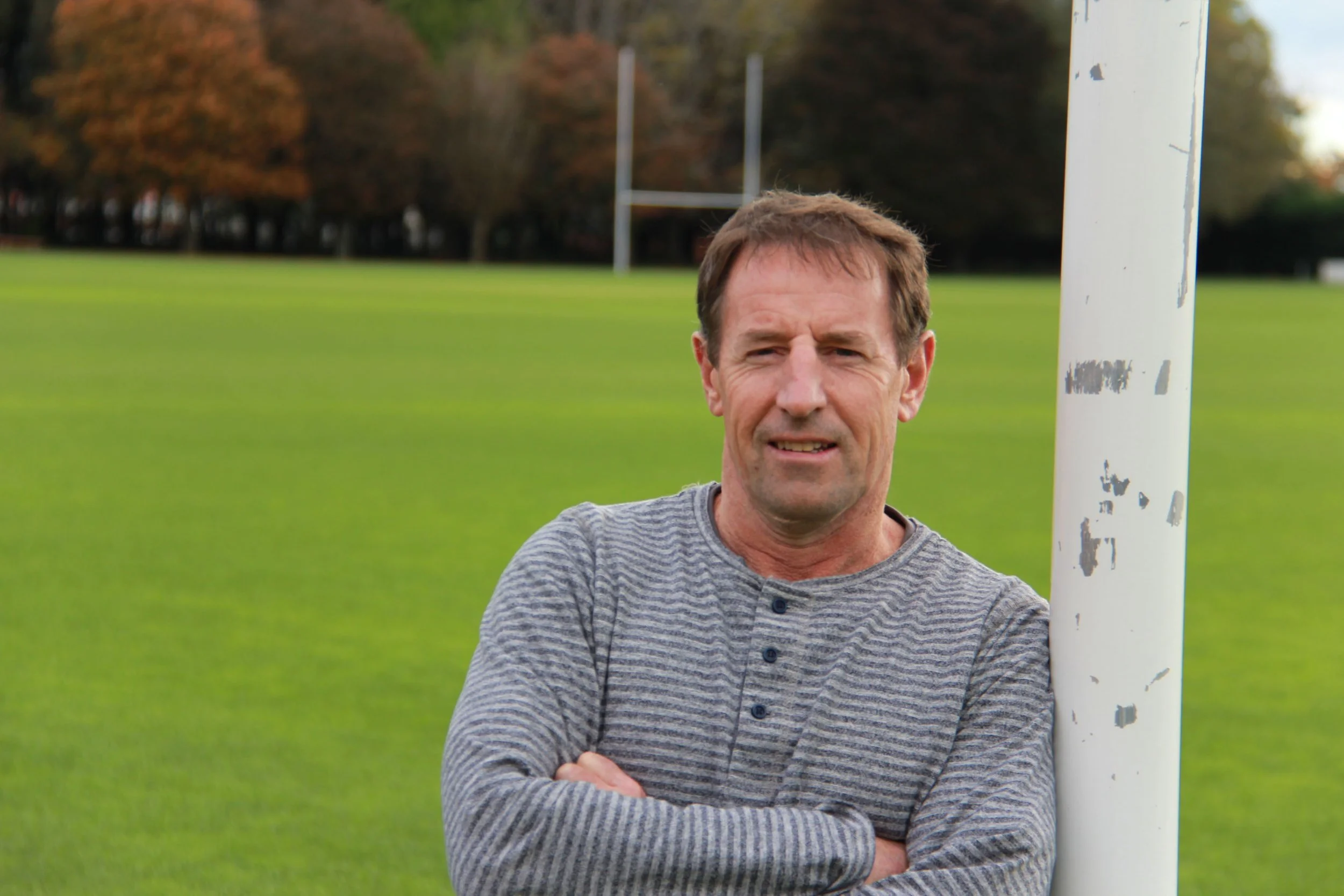 A middle-aged man with brown hair stands outdoors on a grassy field with sports goalposts in the background. He is wearing a grey striped shirt and has his arms crossed, with a serious expression on his face.
