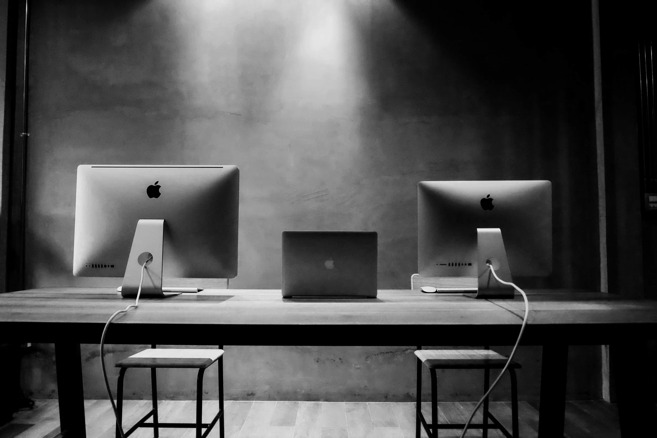 Two Apple iMac computers on a wooden table, with a closed laptop between them, in a minimalist, modern setting with concrete walls and chairs.